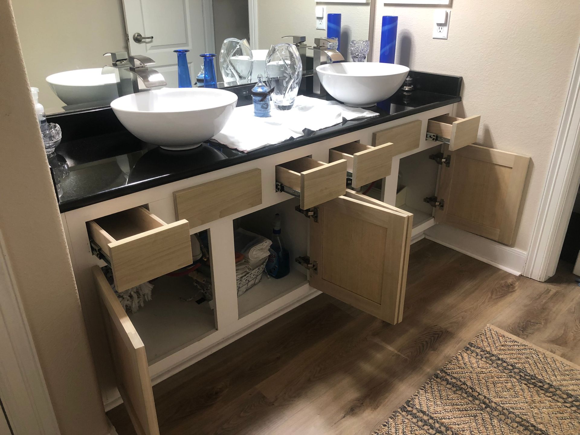 A bathroom vanity with a black countertop and dual white vessel sinks, featuring partially open wooden cabinet drawers.