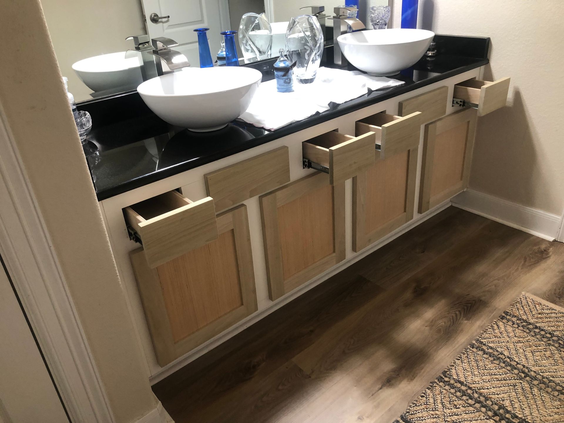 A bathroom vanity with a black countertop and two white vessel sinks, featuring several open, unfinished wooden drawers.