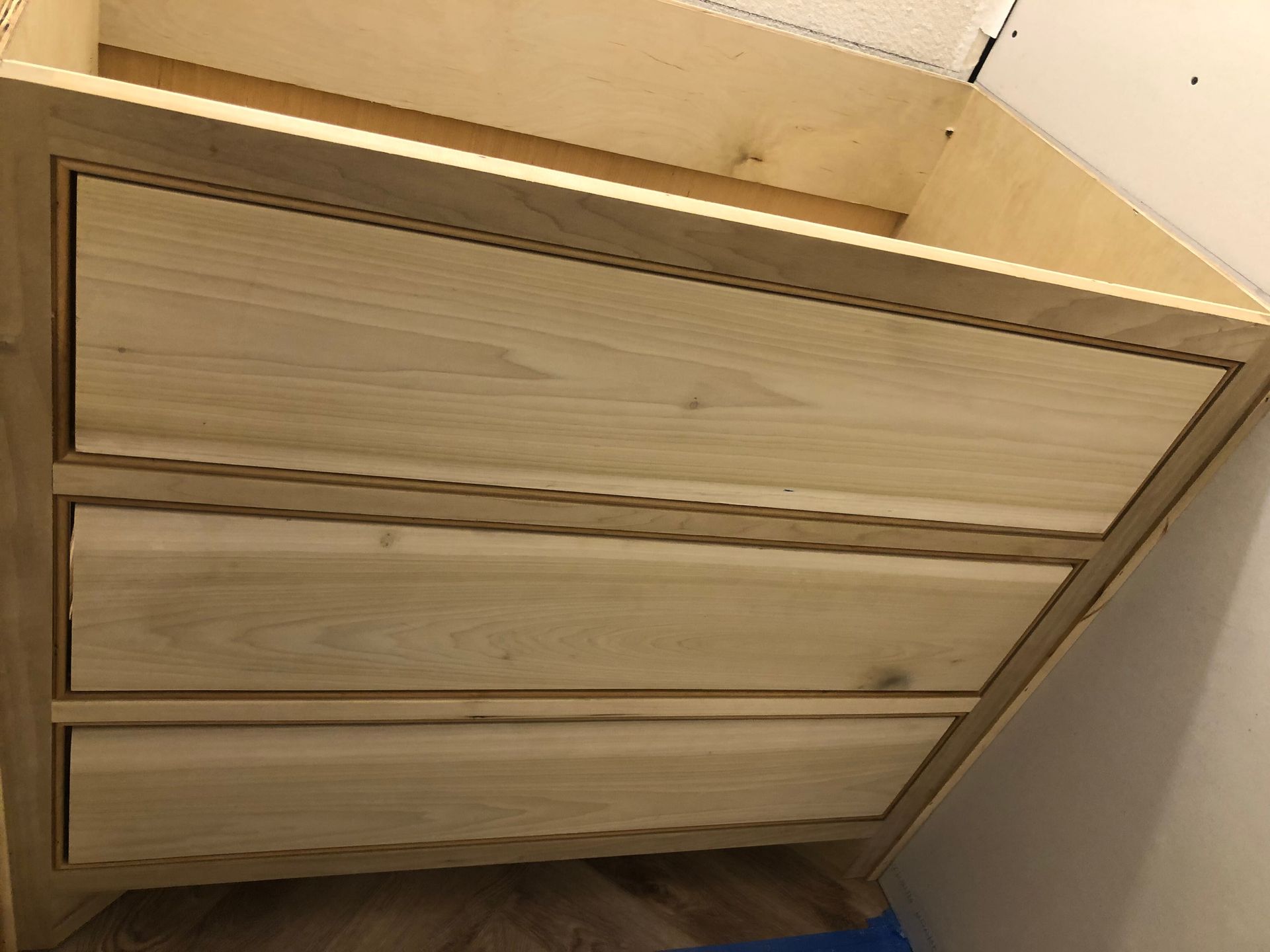 Unfinished light-colored wooden bathroom vanity cabinet with three horizontal drawers, seen from a high angle.