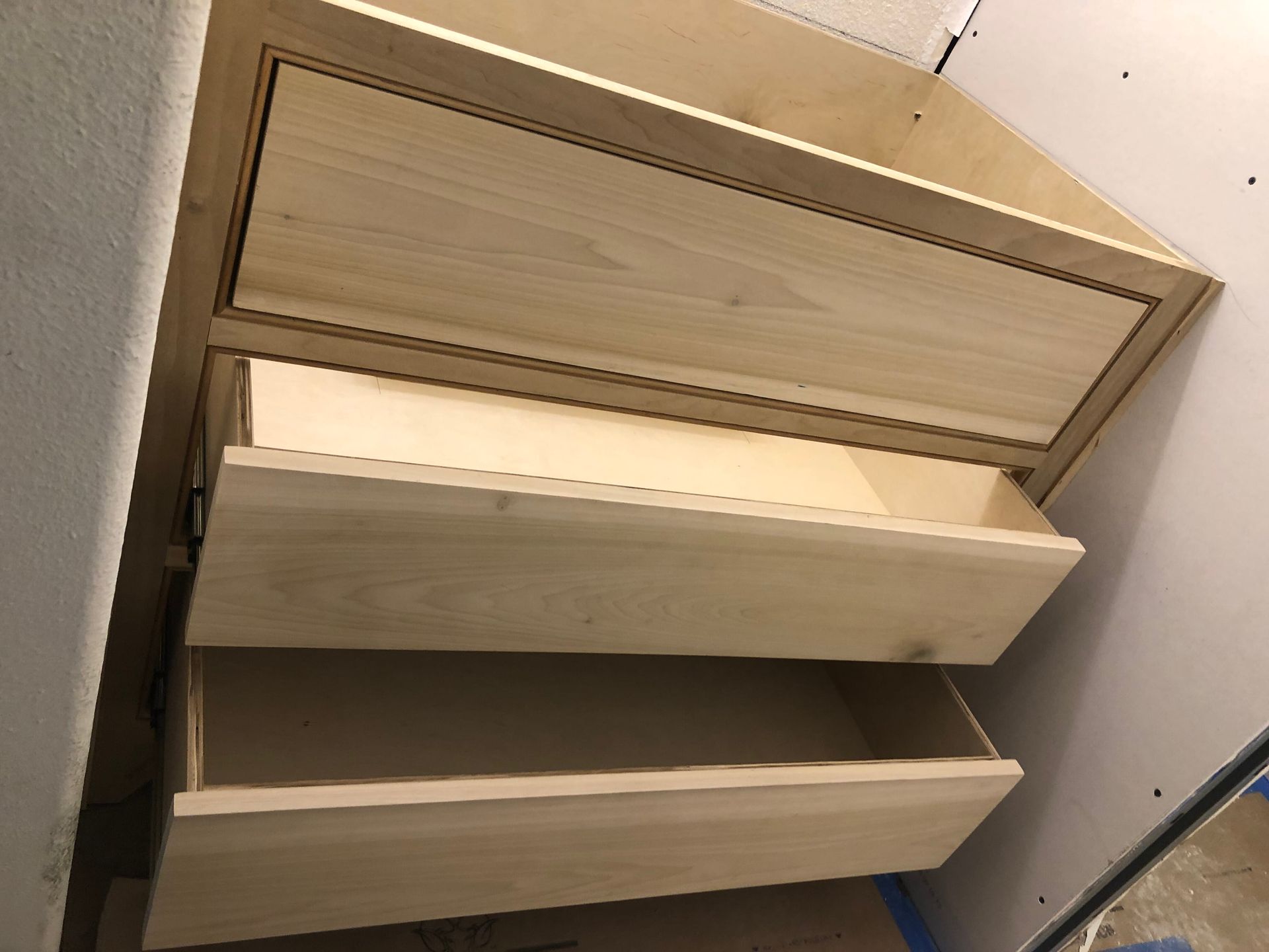 An unfinished, light-colored wooden vanity or dresser with three drawers partially open, set against a wall.