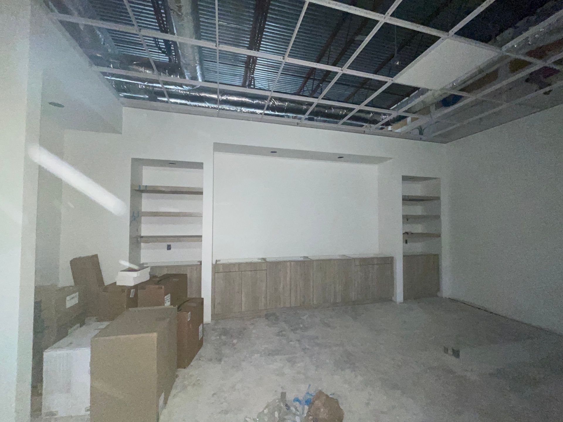 A room under construction with wood-toned base cabinets and built-in shelves against a wall, plus an exposed ceiling grid.