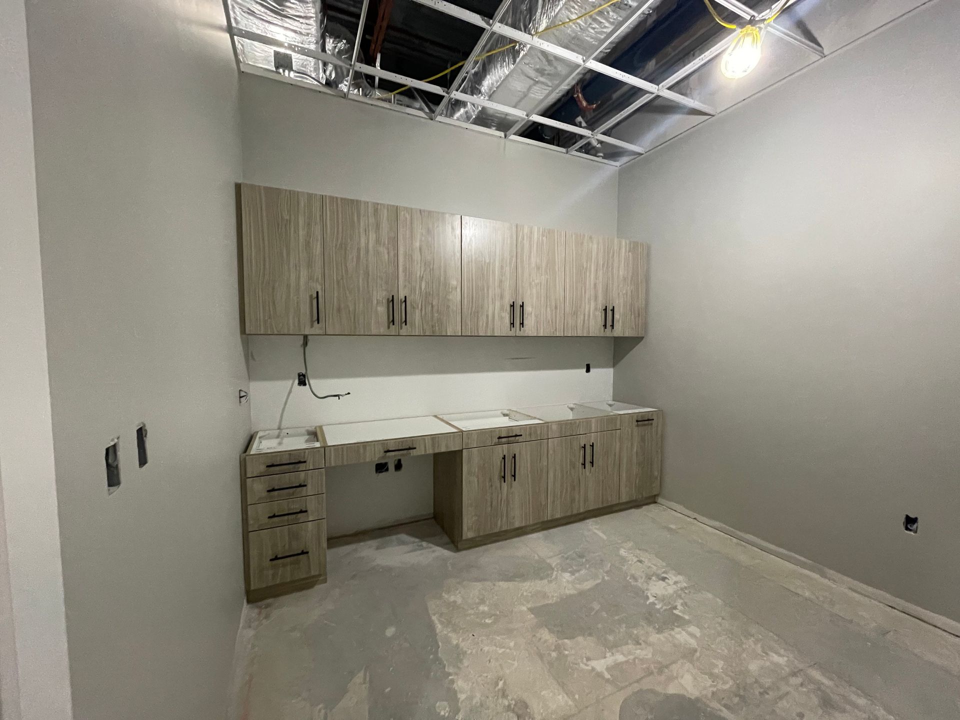 Light wood-toned cabinets installed above a matching workspace and drawers in a room with unfinished gray walls and ceiling.