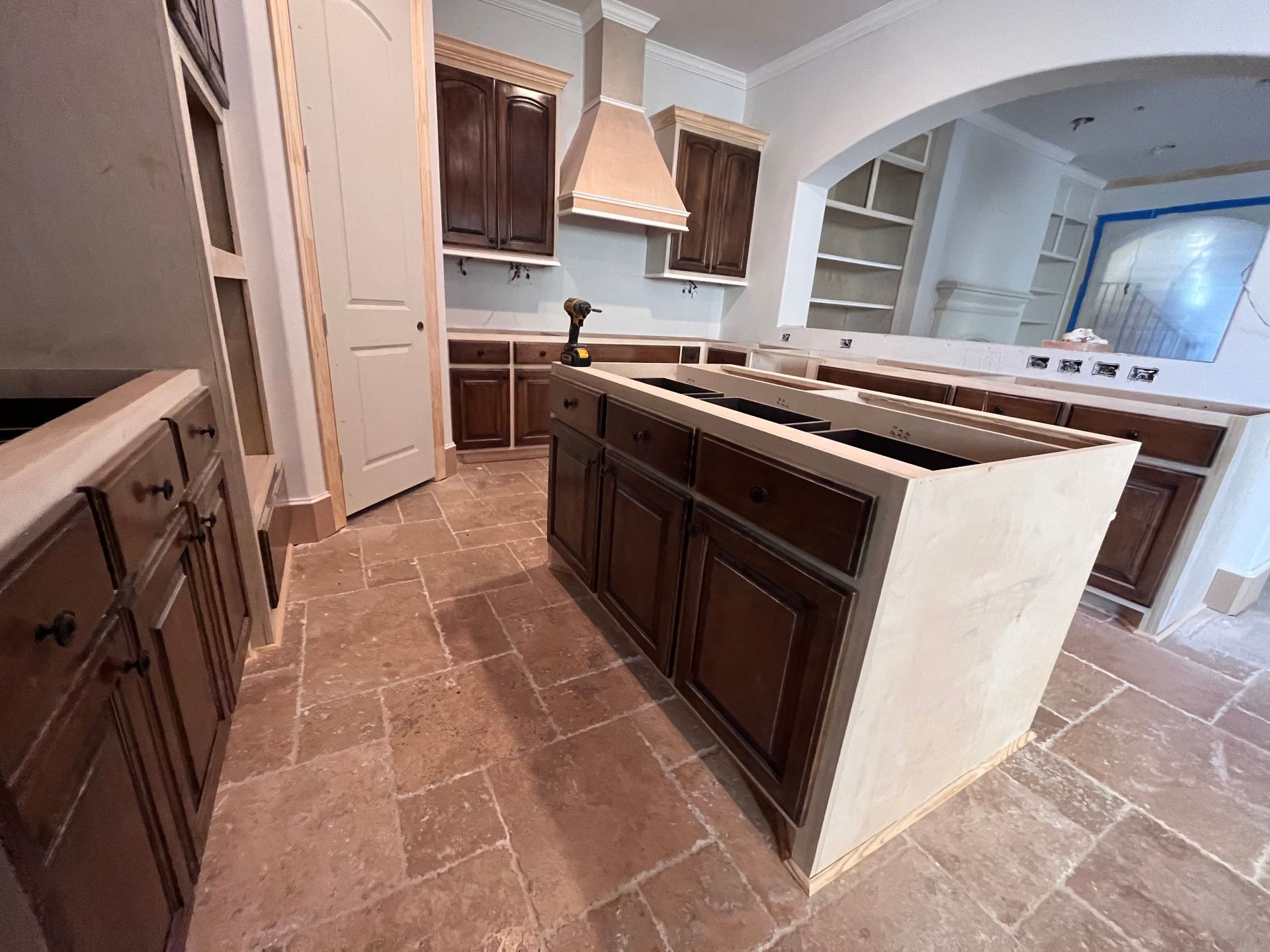 A kitchen under construction featuring dark wood cabinets, a large central island, and brown stone tile flooring.