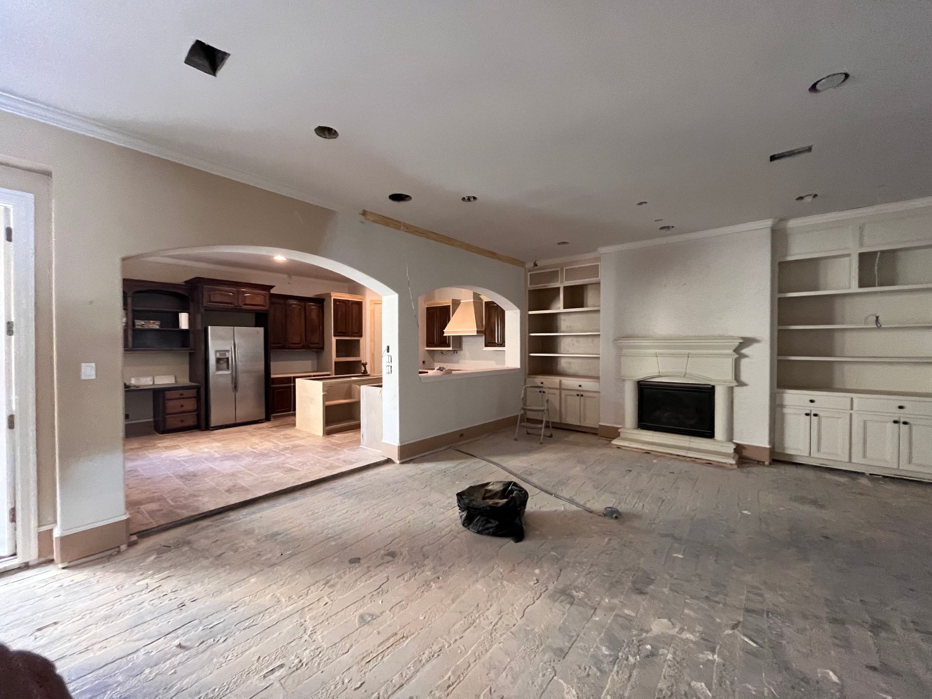 An unfinished, open-plan living room with light walls, built-in shelving, a fireplace, and a view into a kitchen.