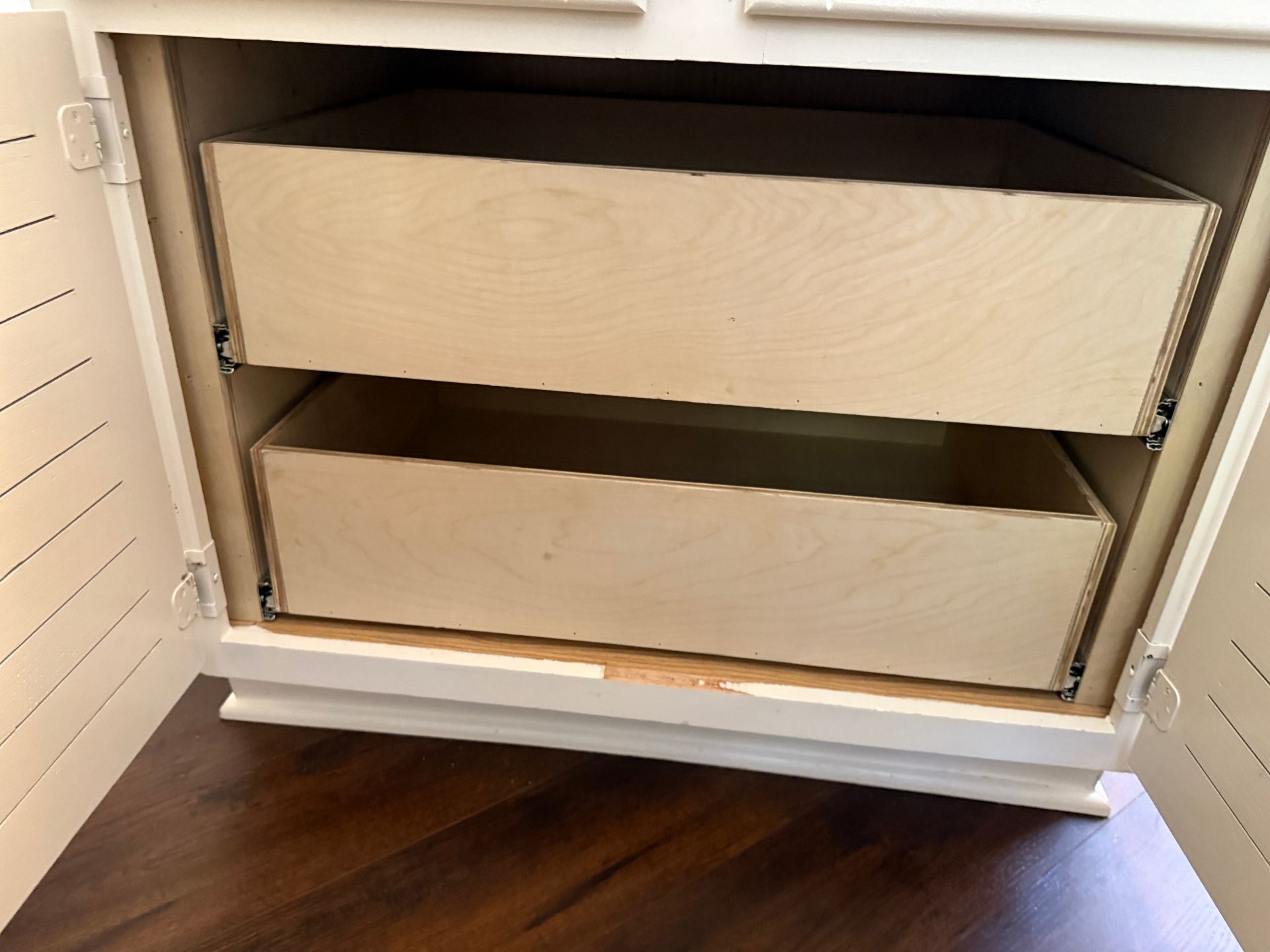 Two light-wood, unfinished drawers inside a white cabinet with slatted doors over a dark wood floor.