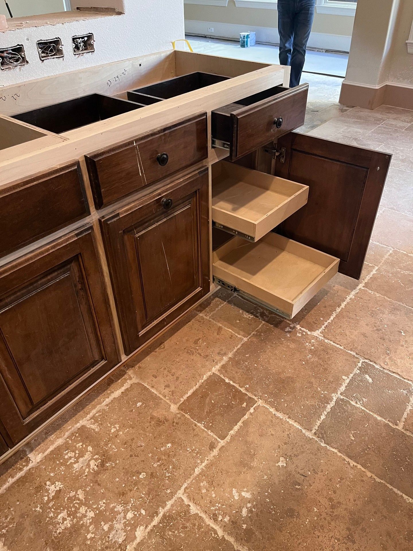 A brown wooden vanity cabinet with an open door and two pull-out shelves, set on a stone tile floor during construction.