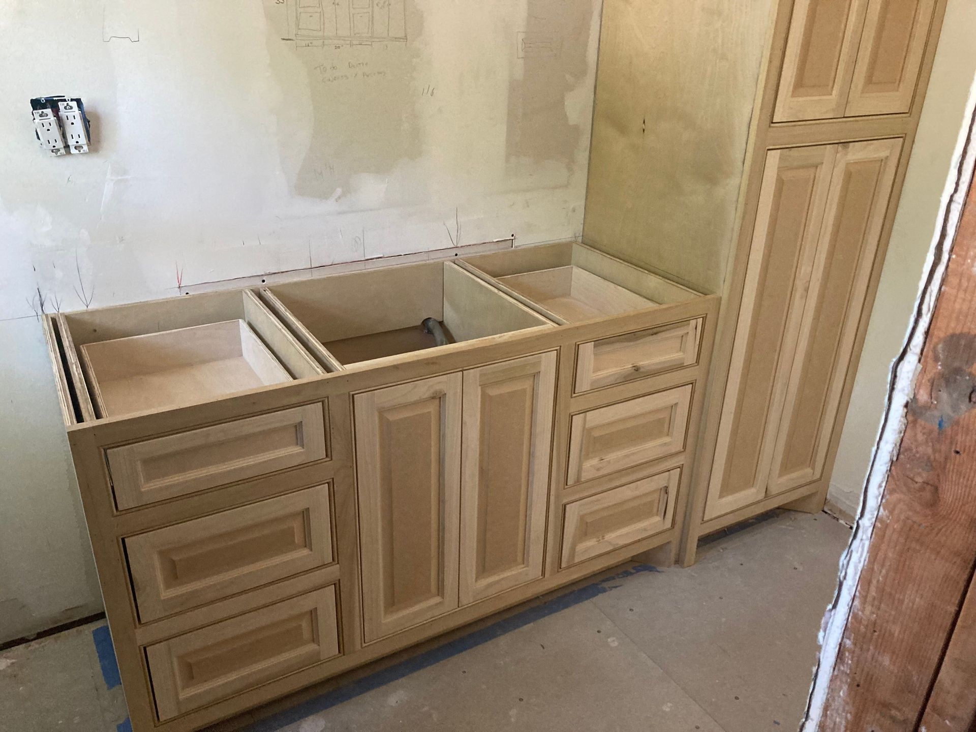 Unfinished light wood bathroom vanity cabinets with drawers and doors installed against a bare wall in a room under remodel.