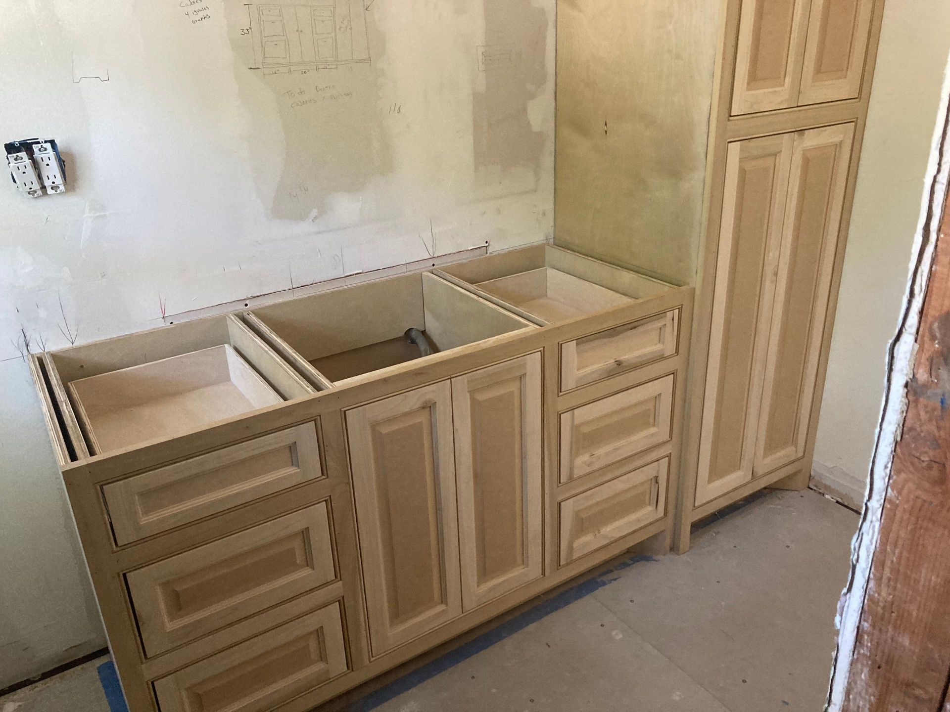 Unfinished light-wood bathroom vanity cabinets with a matching tall storage unit installed against a wall.