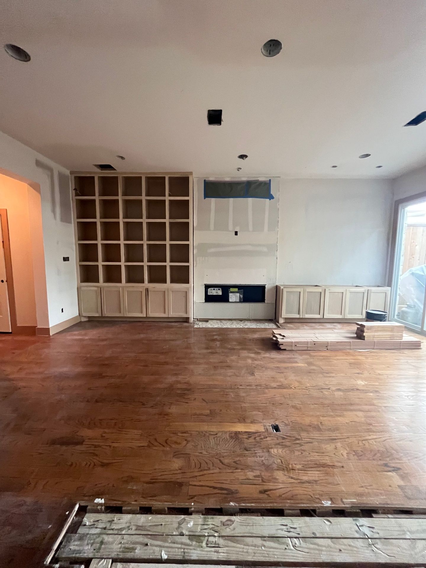 An unfinished room with hardwood floors, a large built-in bookshelf, and a central fireplace area under construction.