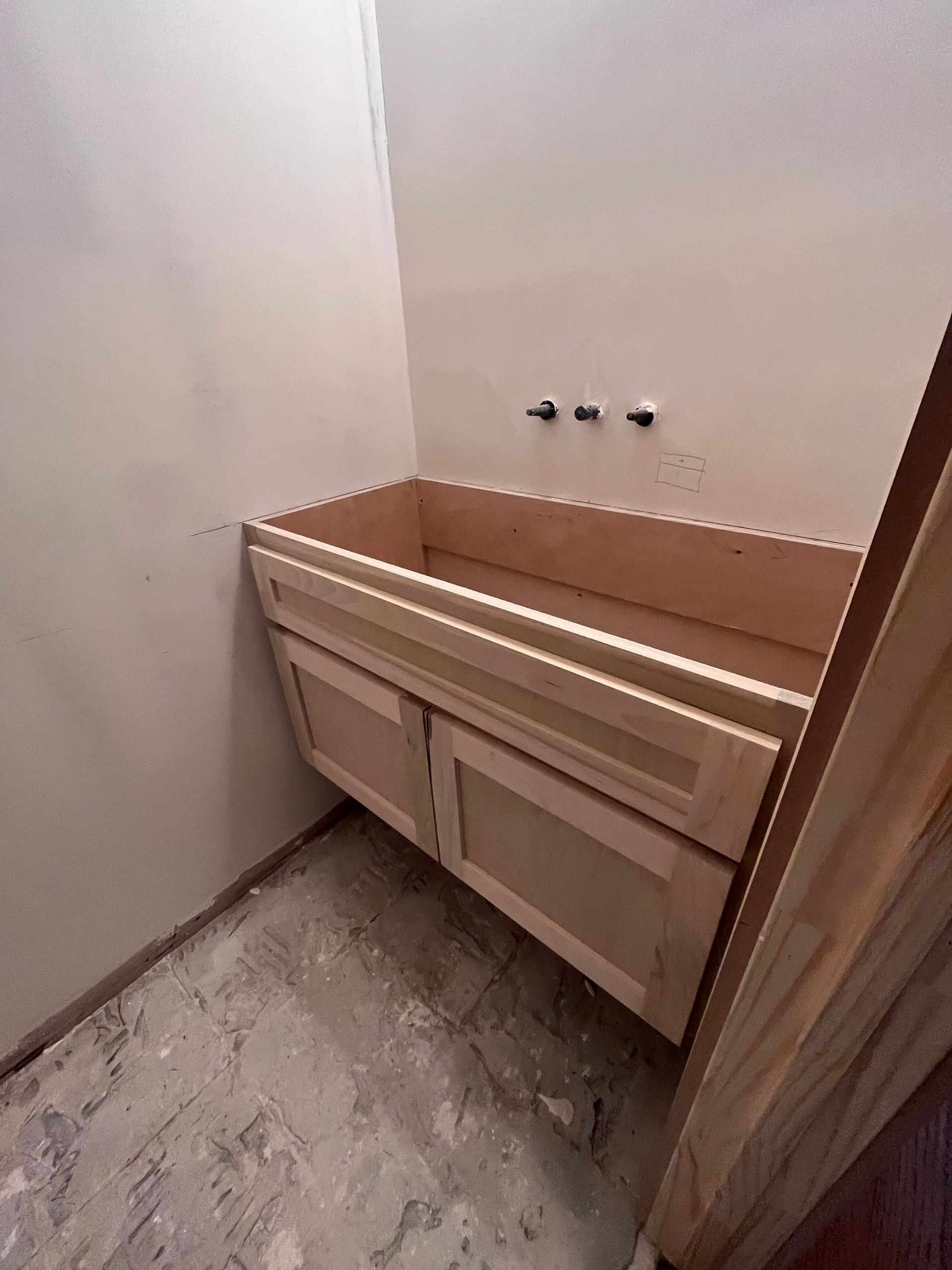 A light-wood bathroom vanity cabinet installed against a white wall with wall-mounted plumbing rough-ins.