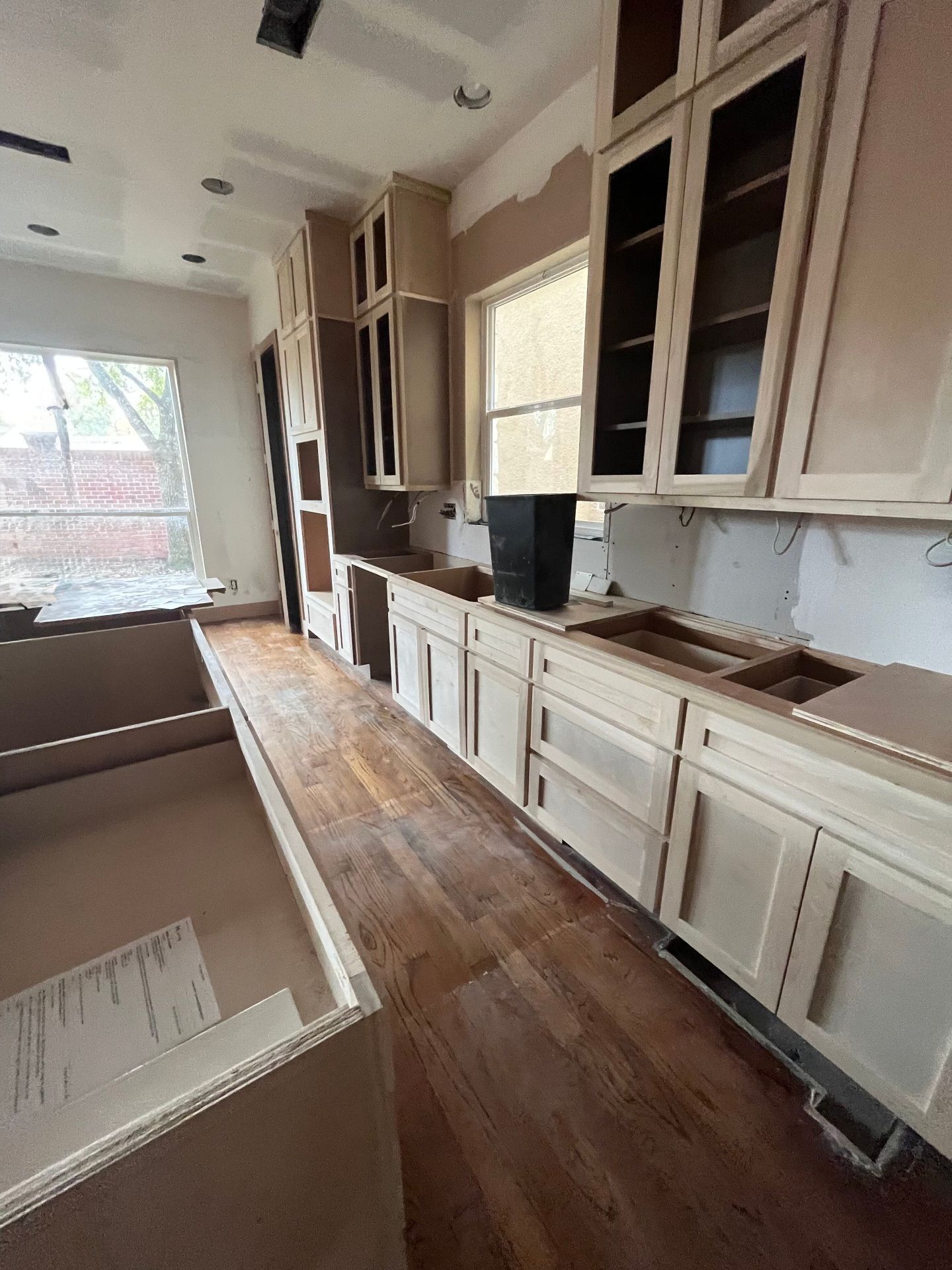 An unfinished kitchen with light wood cabinets, hardwood floors, and a window, currently under renovation.