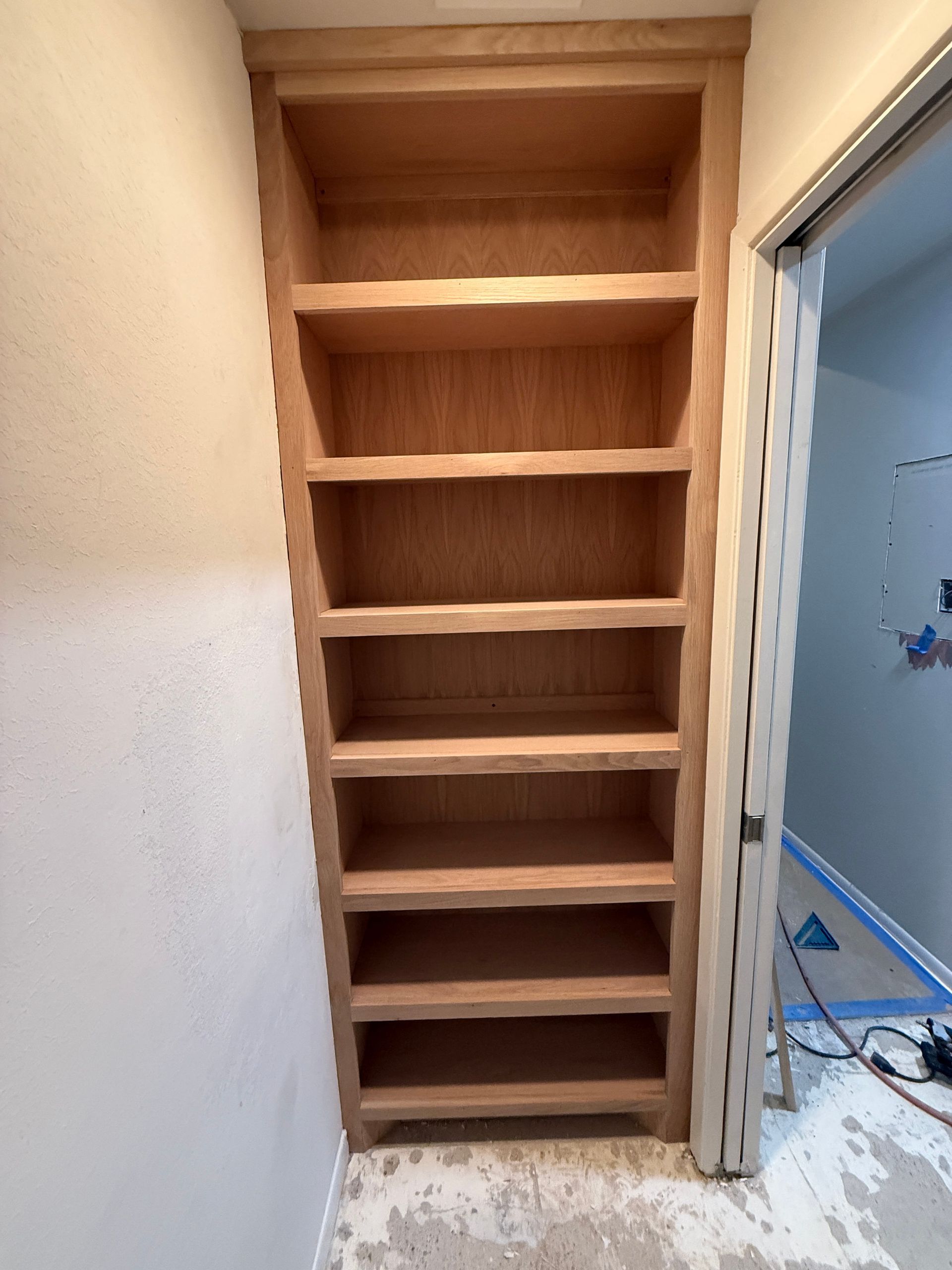 A built-in, unfinished wooden shelving unit with seven shelves, set into a wall next to an open doorway.