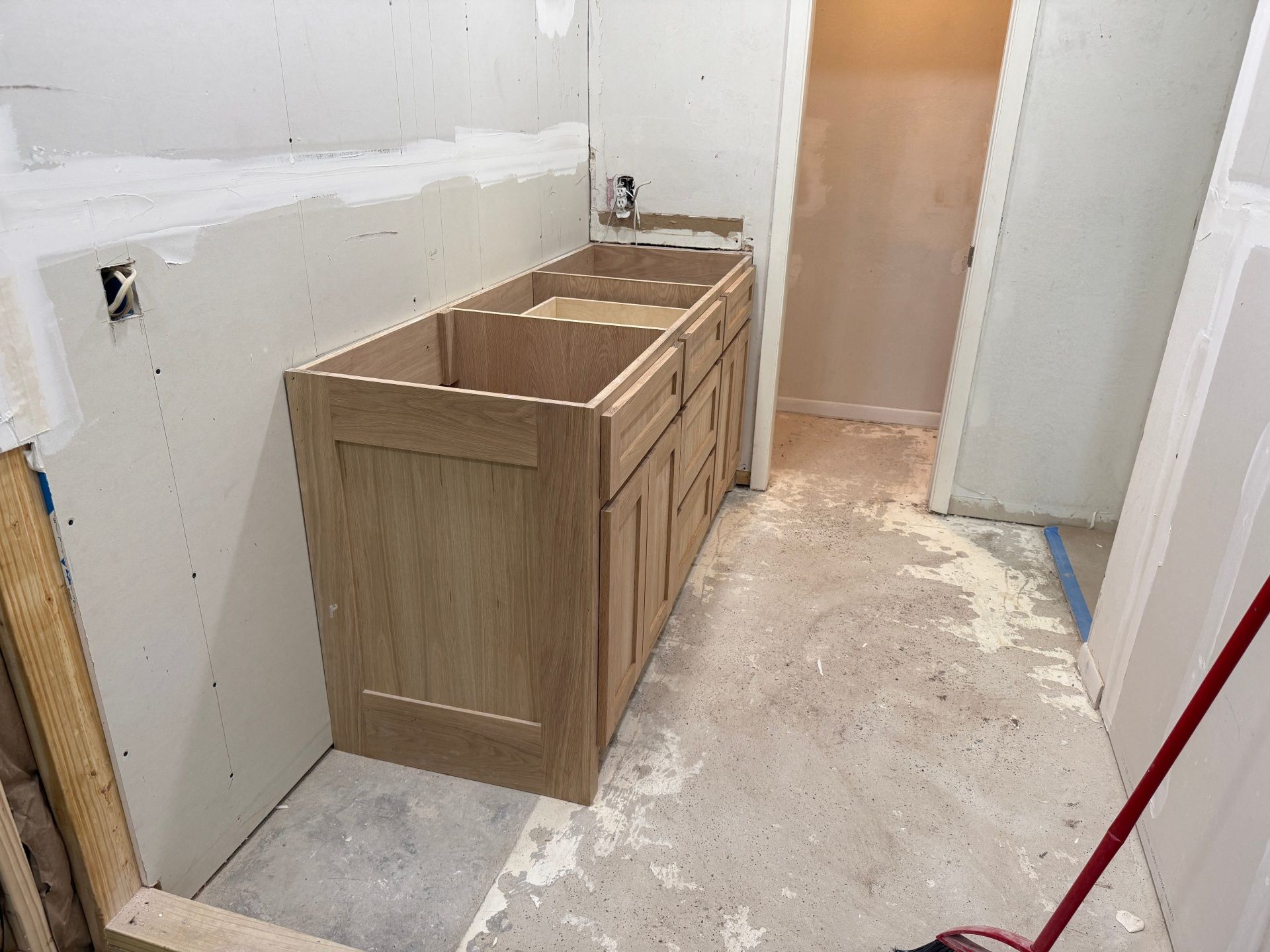 Unfinished light wood vanity cabinet installed in a bathroom renovation with exposed drywall and a dusty concrete floor.