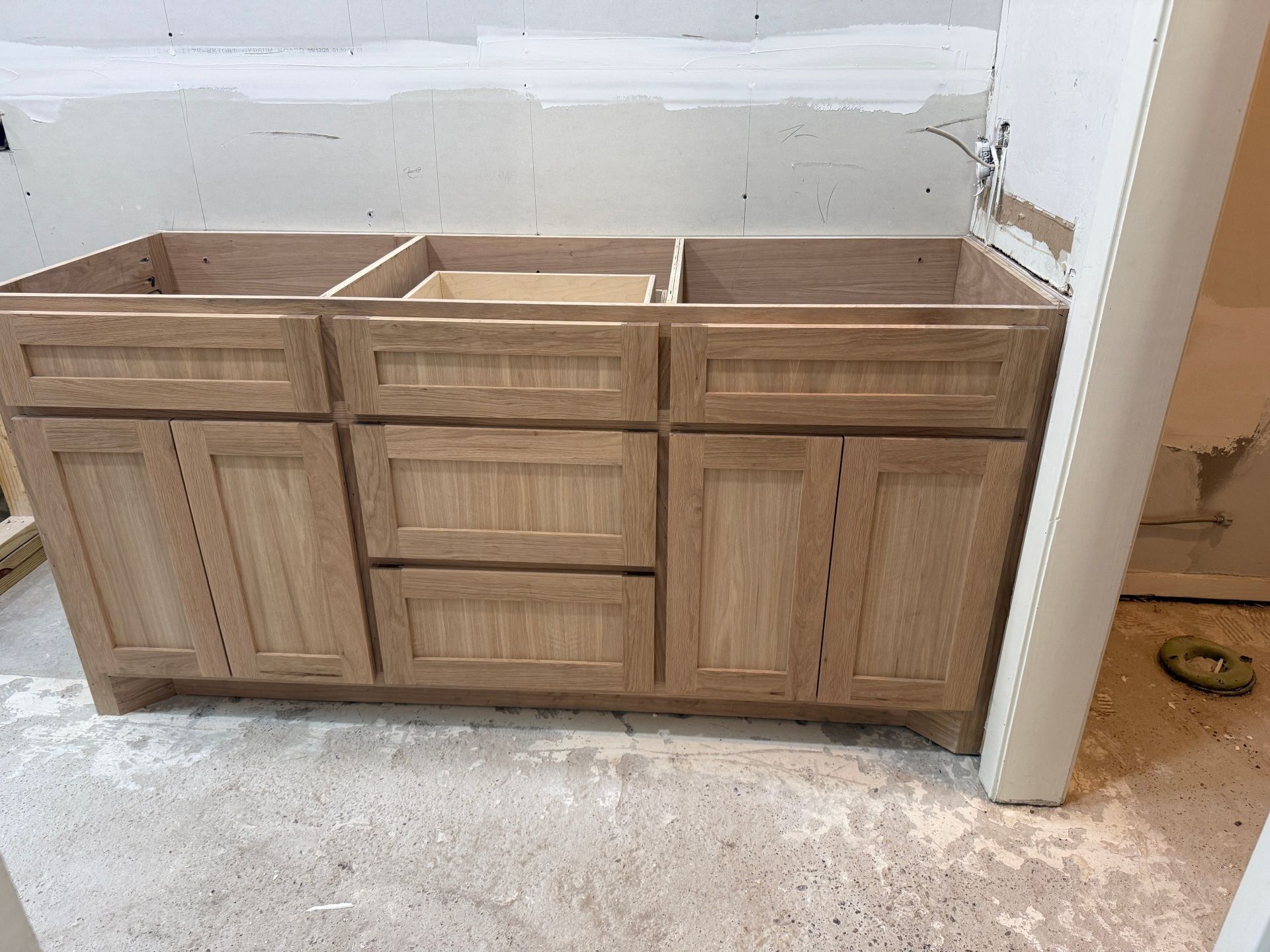 A new, light-wood bathroom vanity cabinet installed against an unfinished drywall wall in a room under construction.