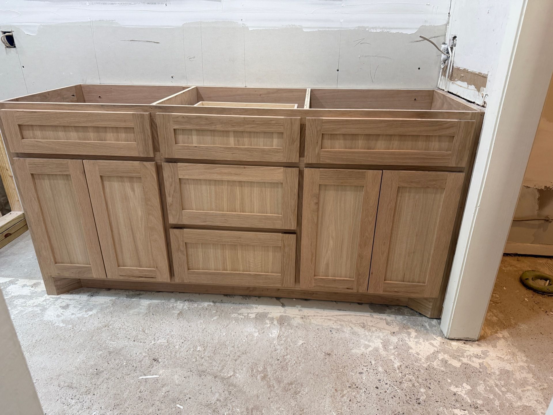 A light wood double bathroom vanity cabinet base installed in a room under construction with unfinished drywall.
