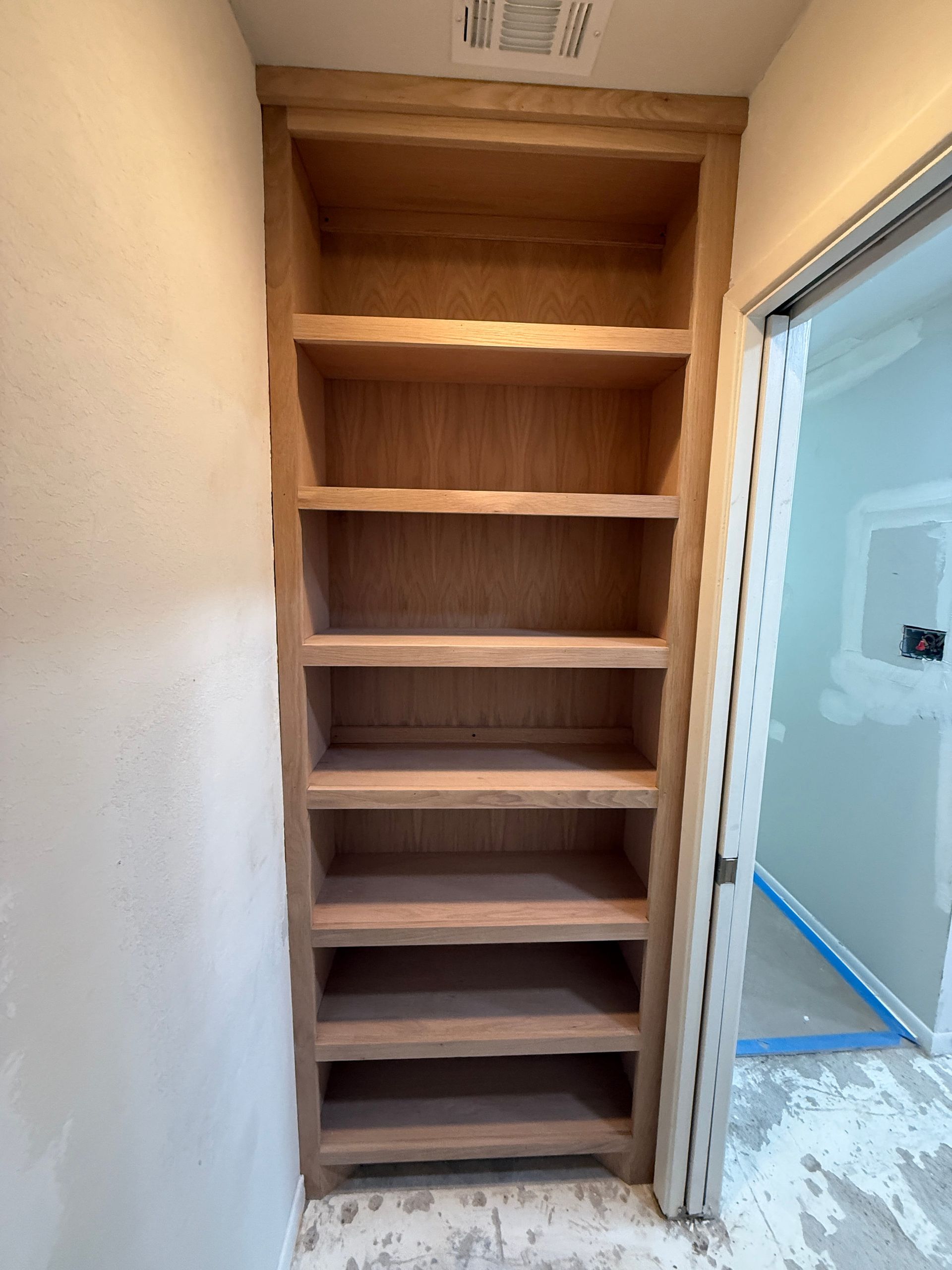 A floor-to-ceiling wooden shelving unit built into an unfinished room with drywall and exposed flooring.