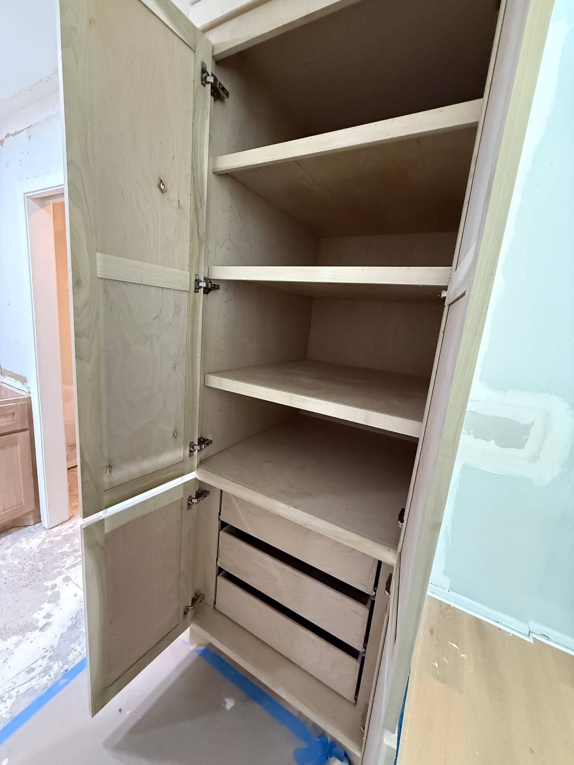 A tall, open-door wooden pantry cabinet with adjustable shelves and three built-in lower drawers in a room under renovation.