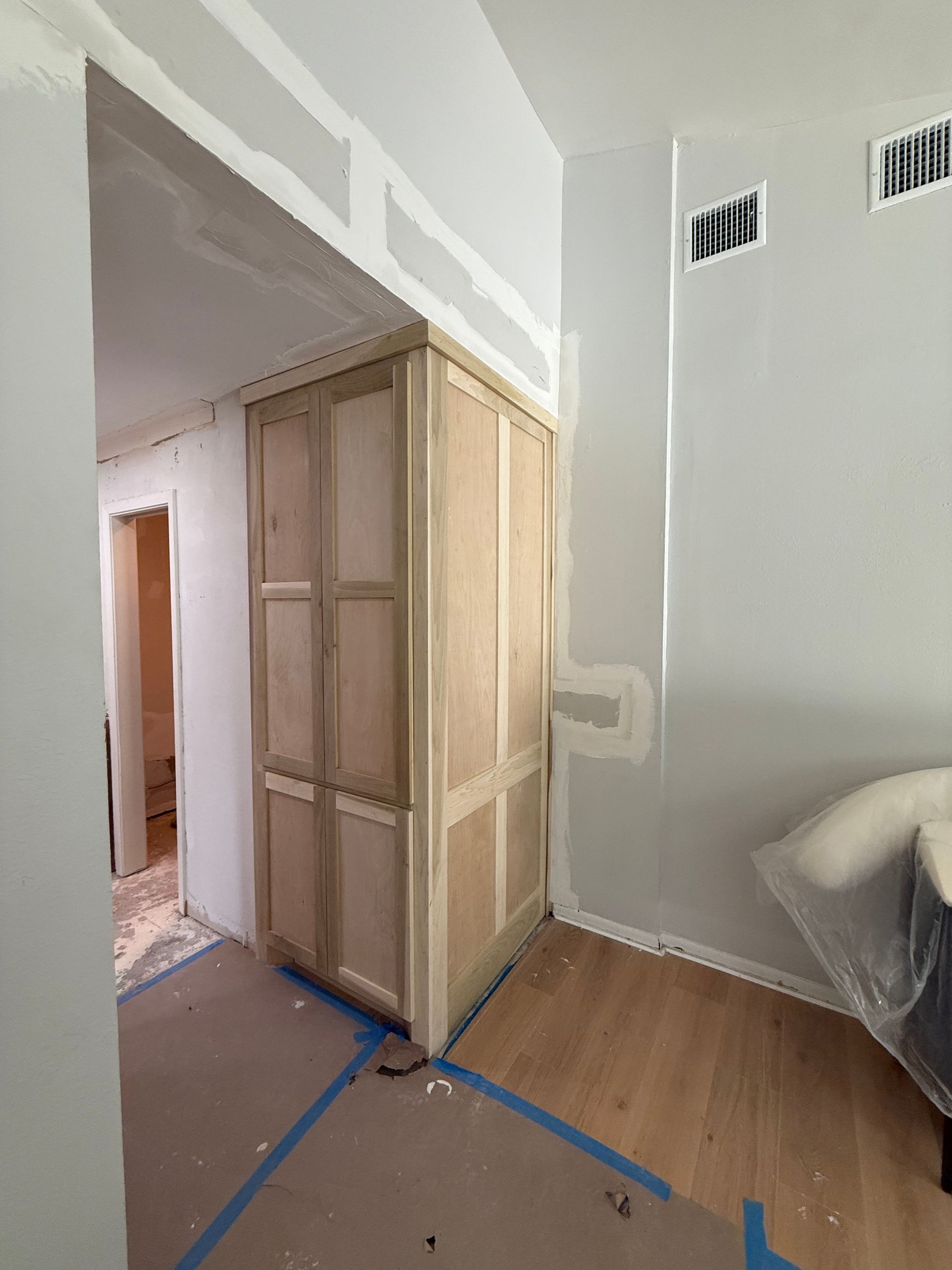 A room interior under renovation with unfinished wood cabinets, drywall patches, and protective paper on the floor.