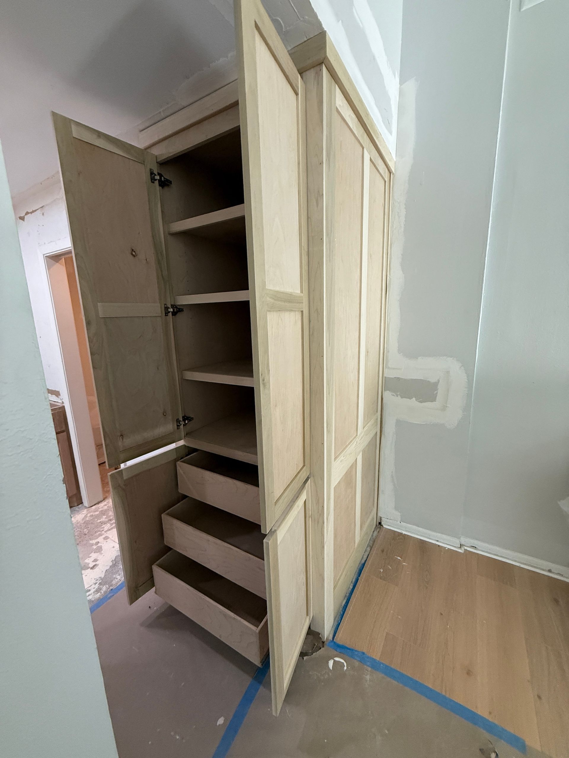 A tall, light-wood pantry cabinet under construction in a room, with the doors open to reveal interior shelves and drawers.