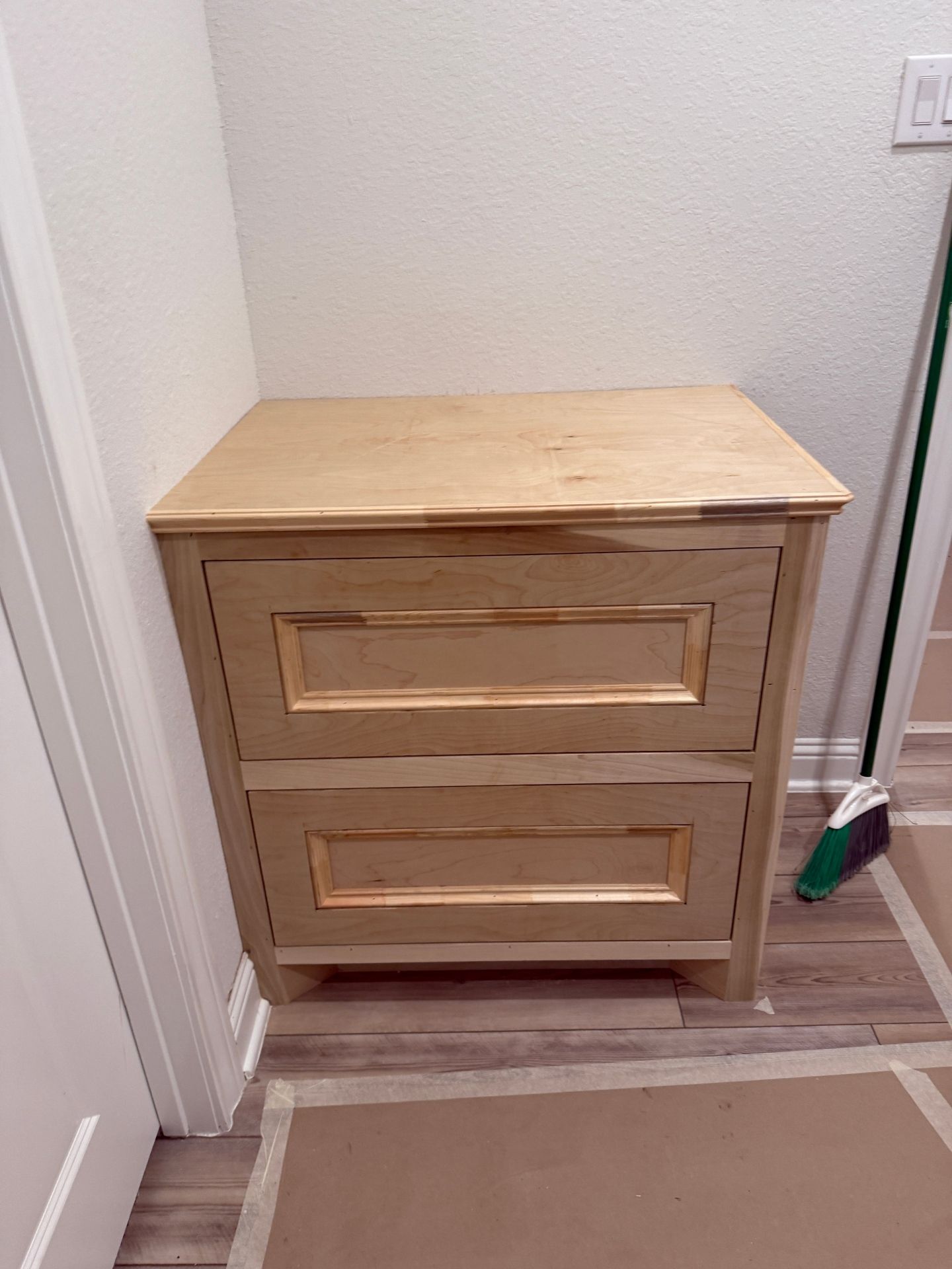 A light wood, two-drawer nightstand with a frame-and-panel design sitting on a wood-look floor against a white wall.