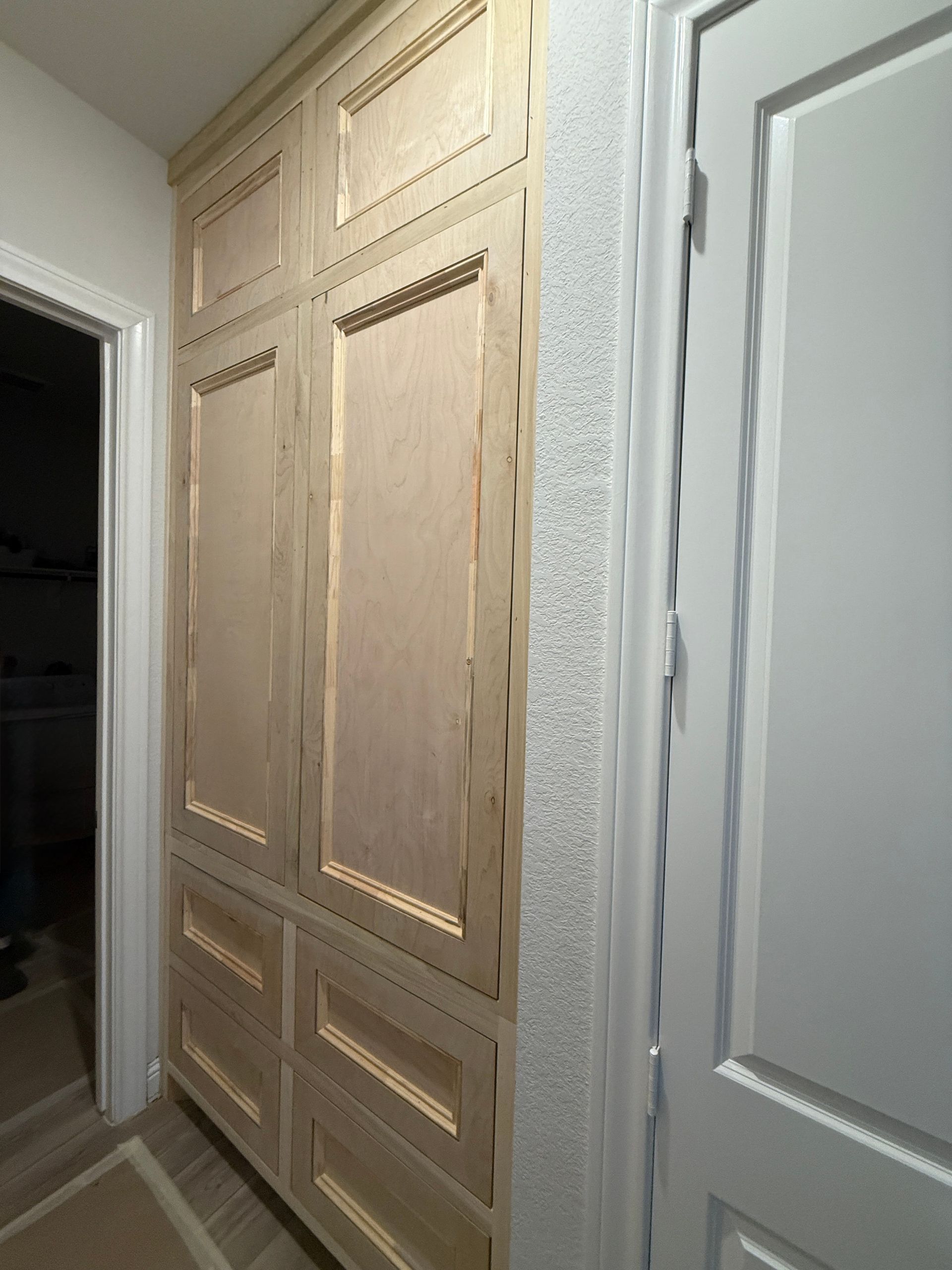 Unfinished wooden built-in cabinets with decorative molding and drawers set in a hallway next to a white door.