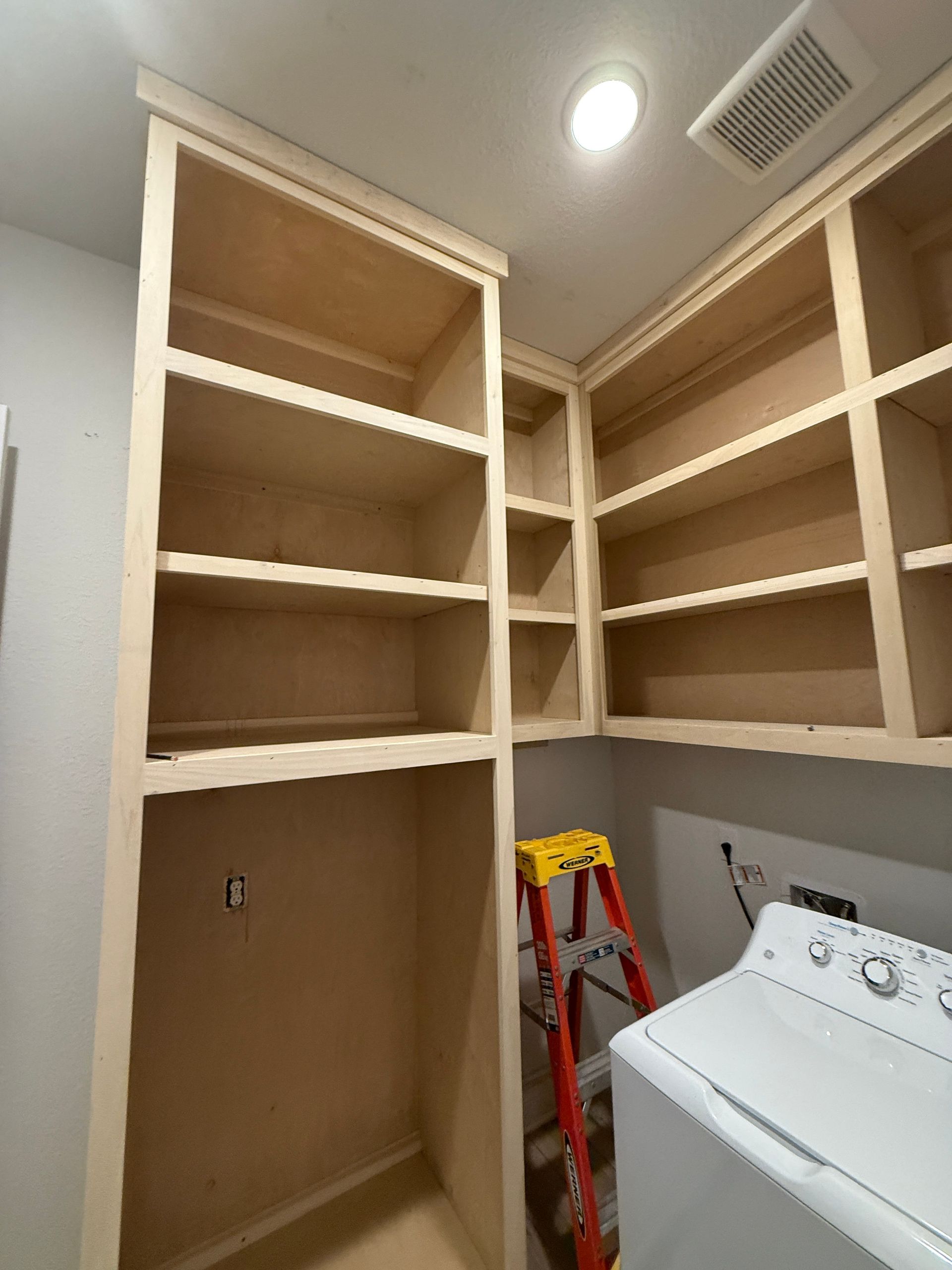 Unfinished wooden shelving units installed on a wall above a washing machine in a laundry room with a ladder nearby.