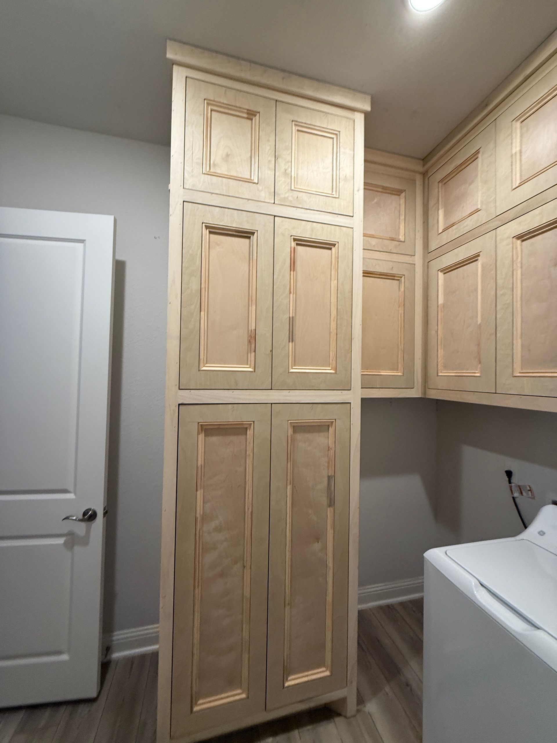 Tall light-colored wooden cabinets installed in a laundry room next to a white door and a washing machine.