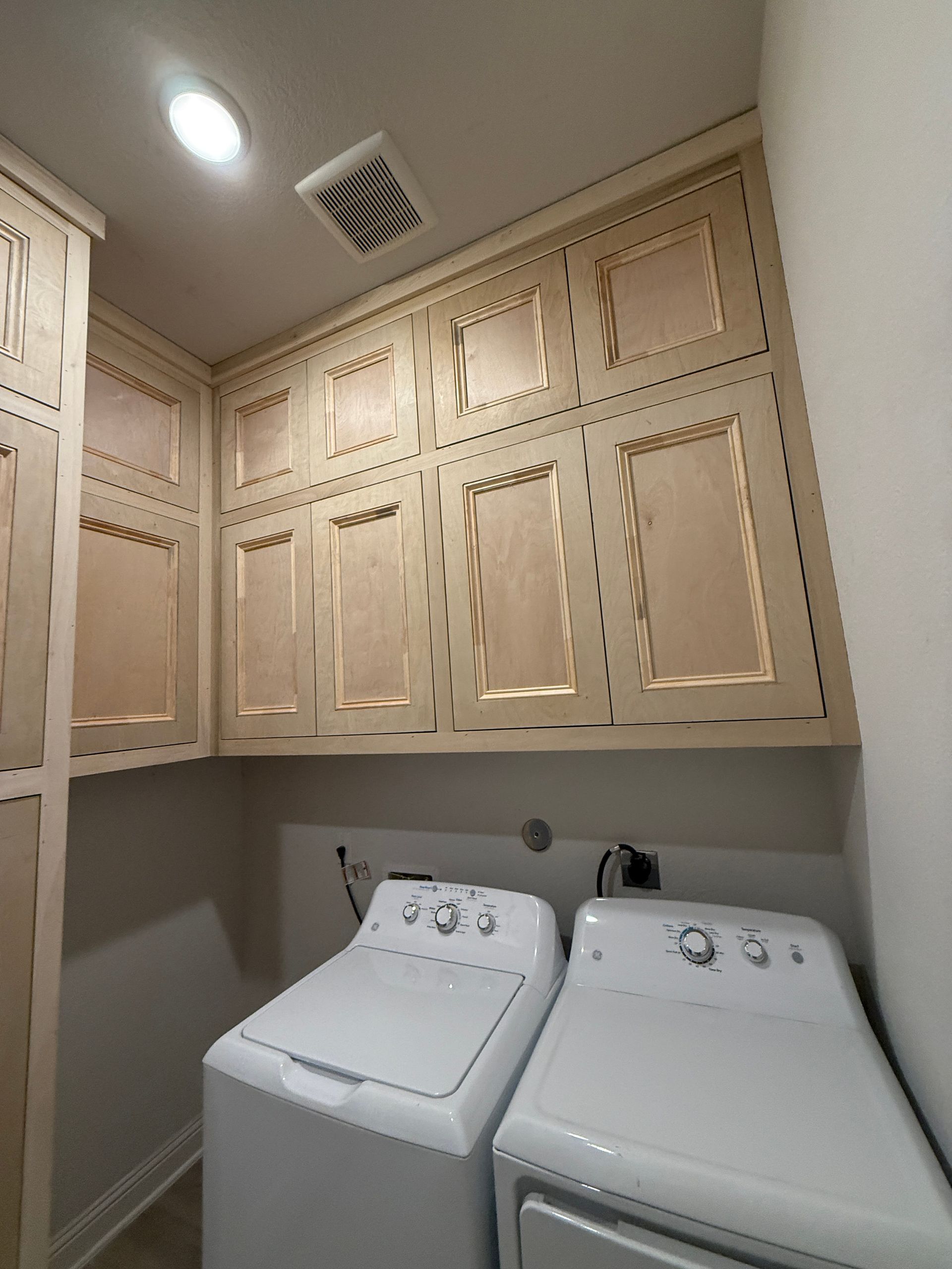 A white washer and dryer sit under beige upper cabinets in a laundry room with a recessed light and ceiling vent.