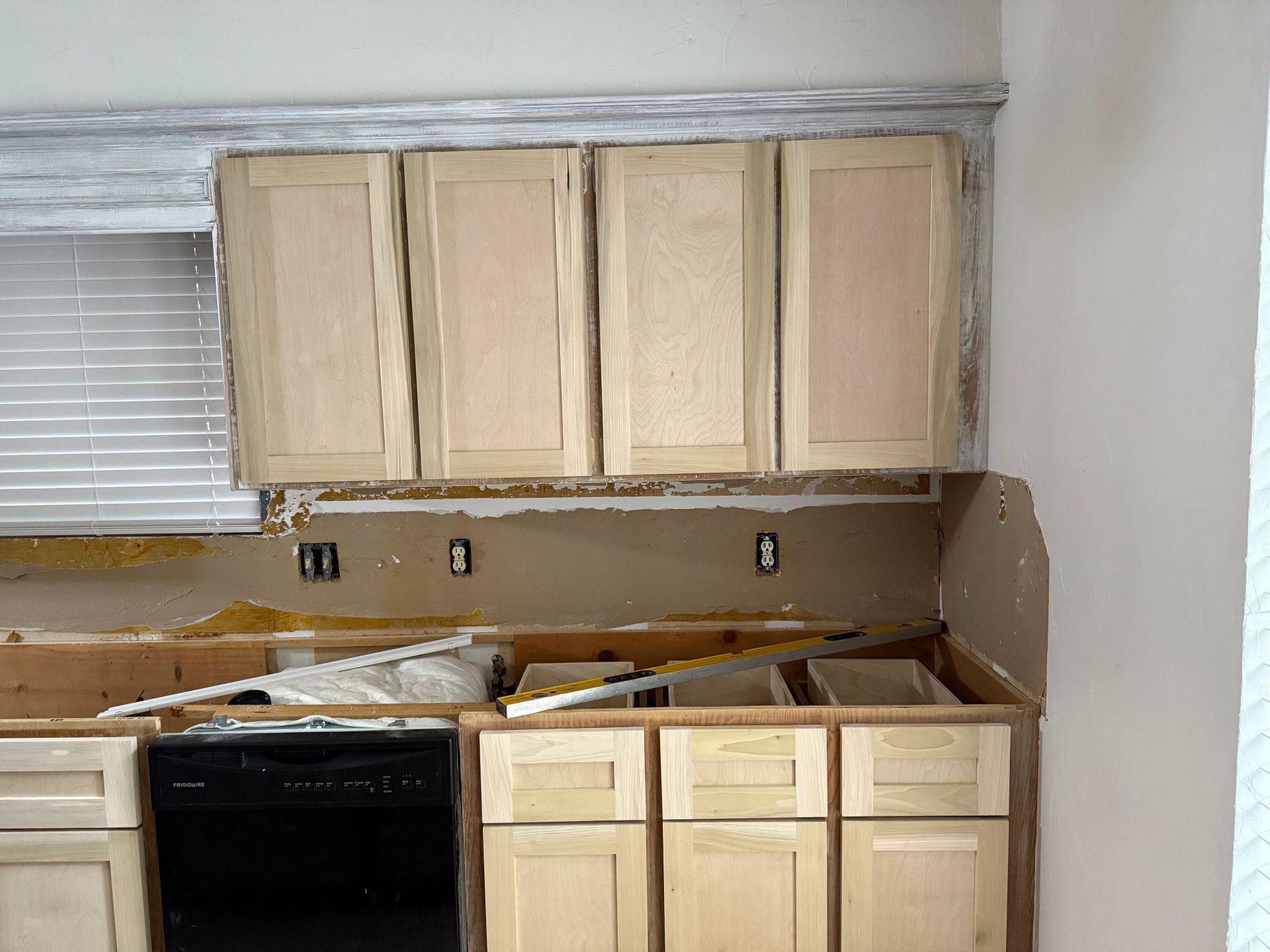 A kitchen renovation in progress featuring unfinished light-wood cabinets, exposed drywall, and a black dishwasher.