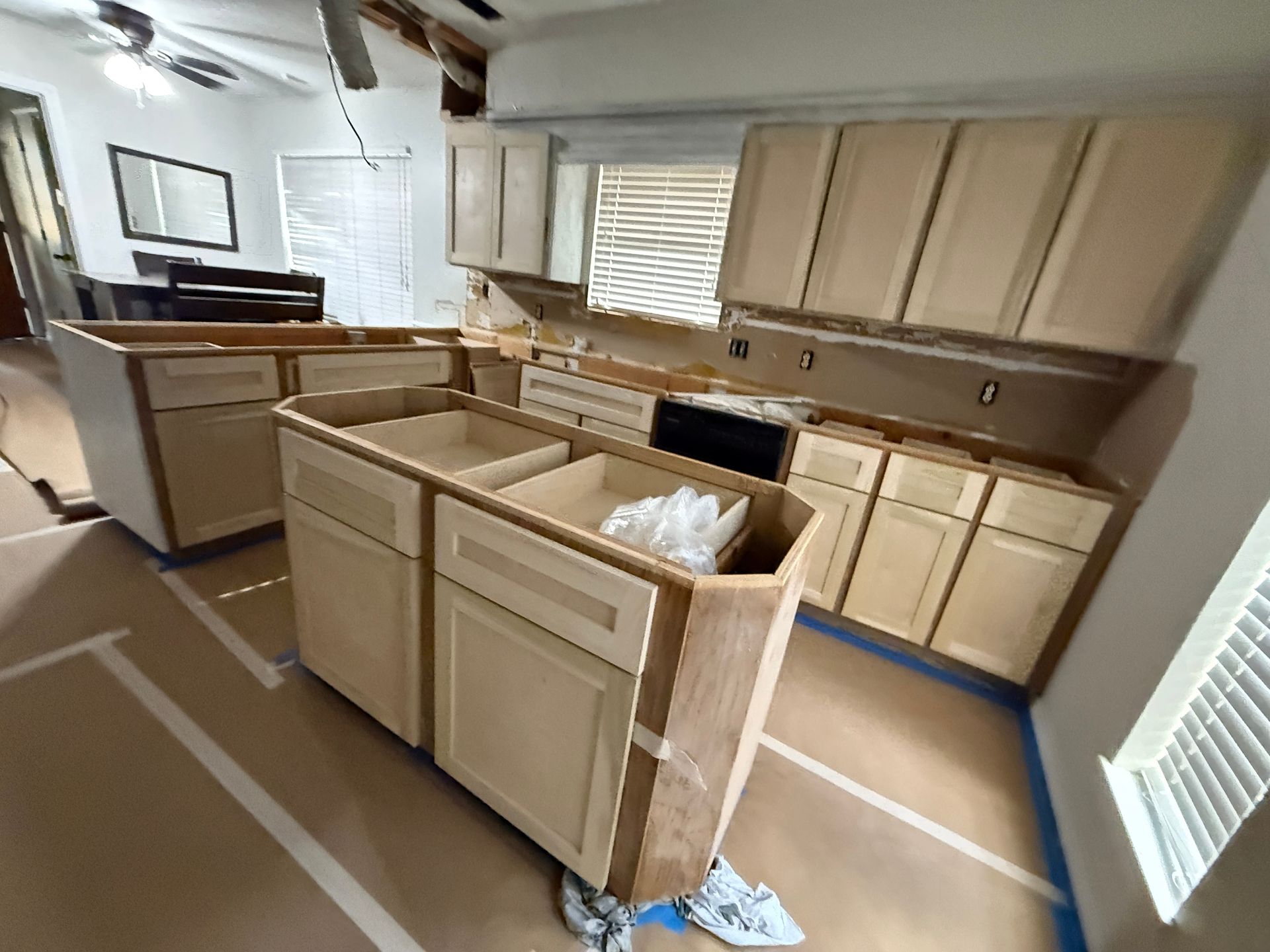 A kitchen under renovation with unfinished, light wood cabinets, an island base, and protective paper on the floor.