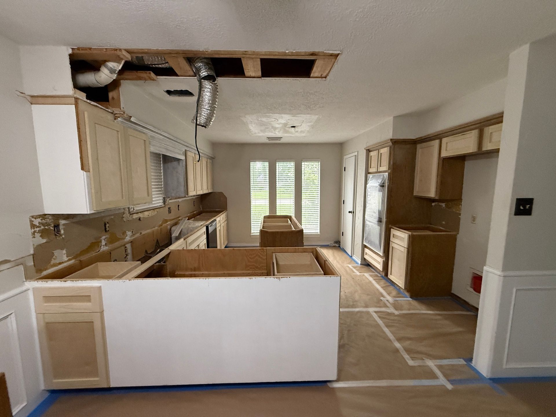 A kitchen under renovation with unfinished wooden cabinets, exposed ductwork, and floor protection paper.