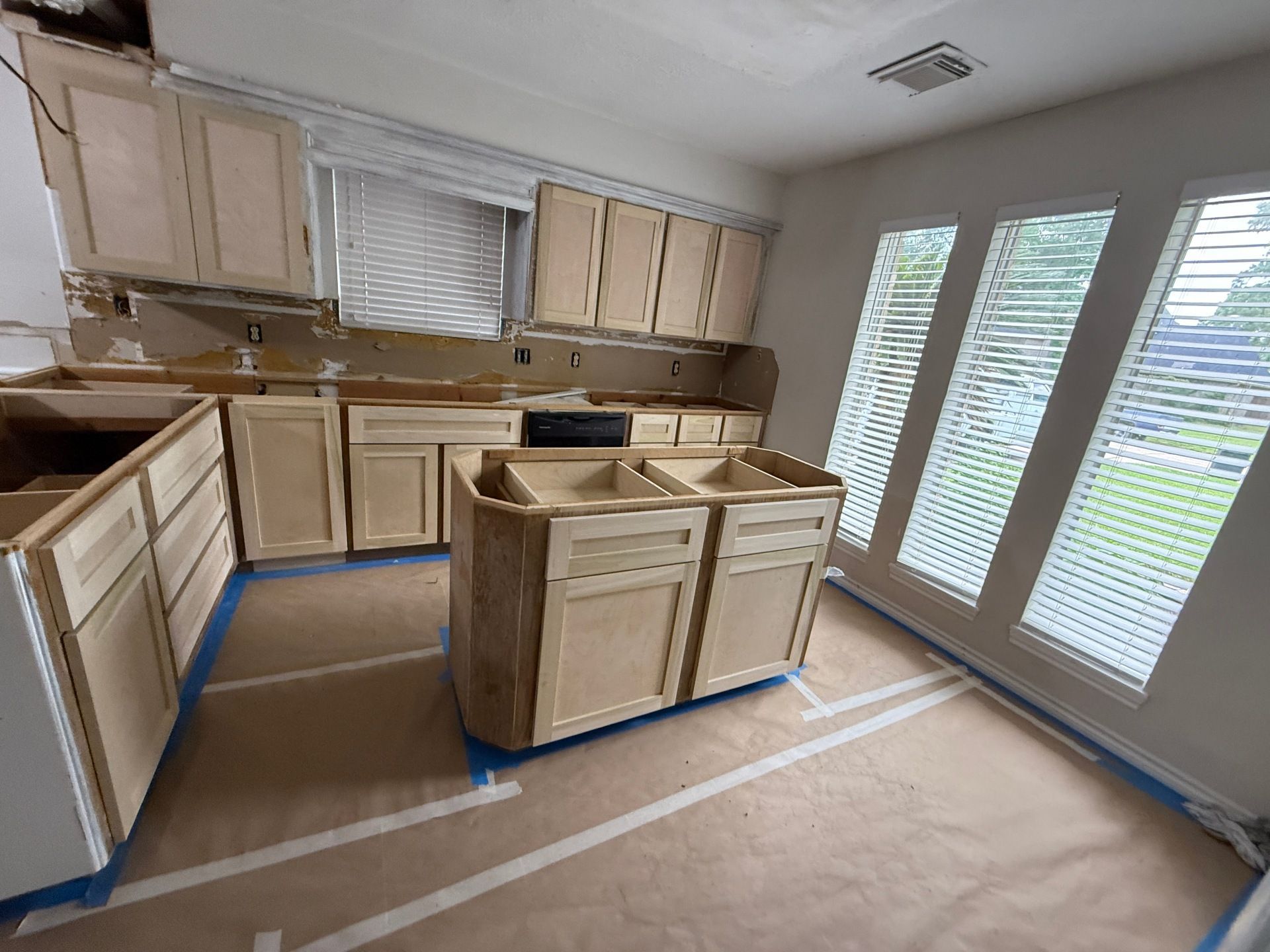A kitchen under construction with unpainted wooden cabinets installed, brown paper on the floor, and three windows.