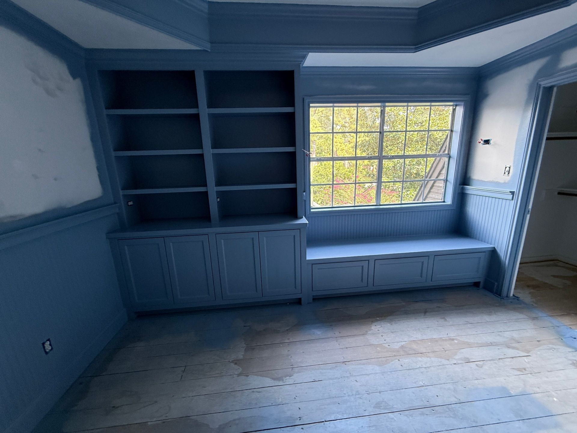 A room under renovation with dusty floors, featuring built-in blue shelving and a window seat painted in matching blue.