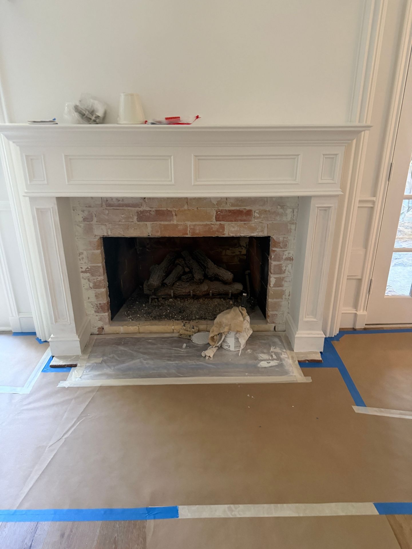 A white fireplace mantel surrounds a brick firebox opening, with paper floor protection and blue painter's tape below.