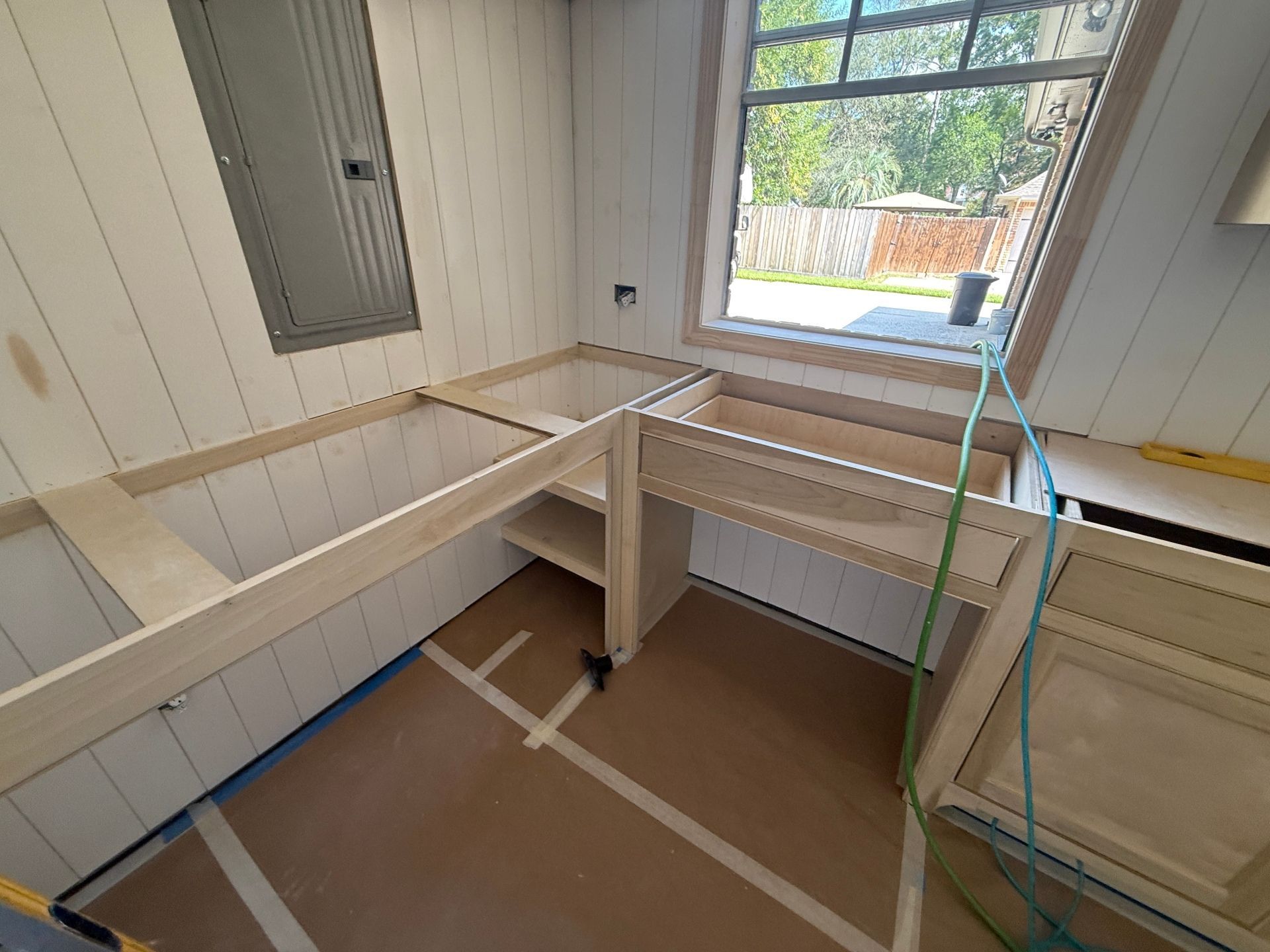 Unfinished light-colored wooden kitchen cabinets frame a corner with a window, set against white vertical panel walls.