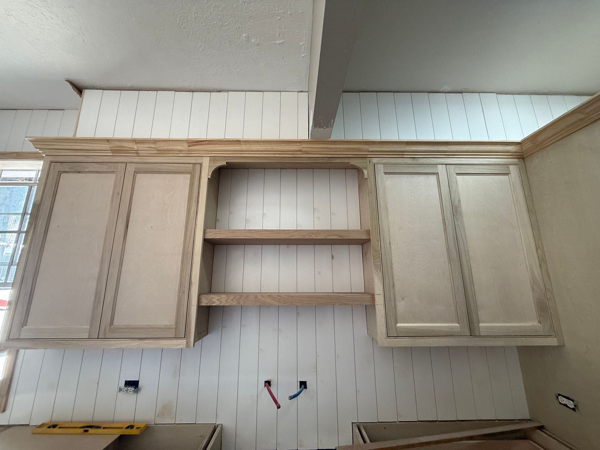 Kitchen cabinets in progress with crown molding, open shelving in the center, and unfinished light wood against a backsplash.