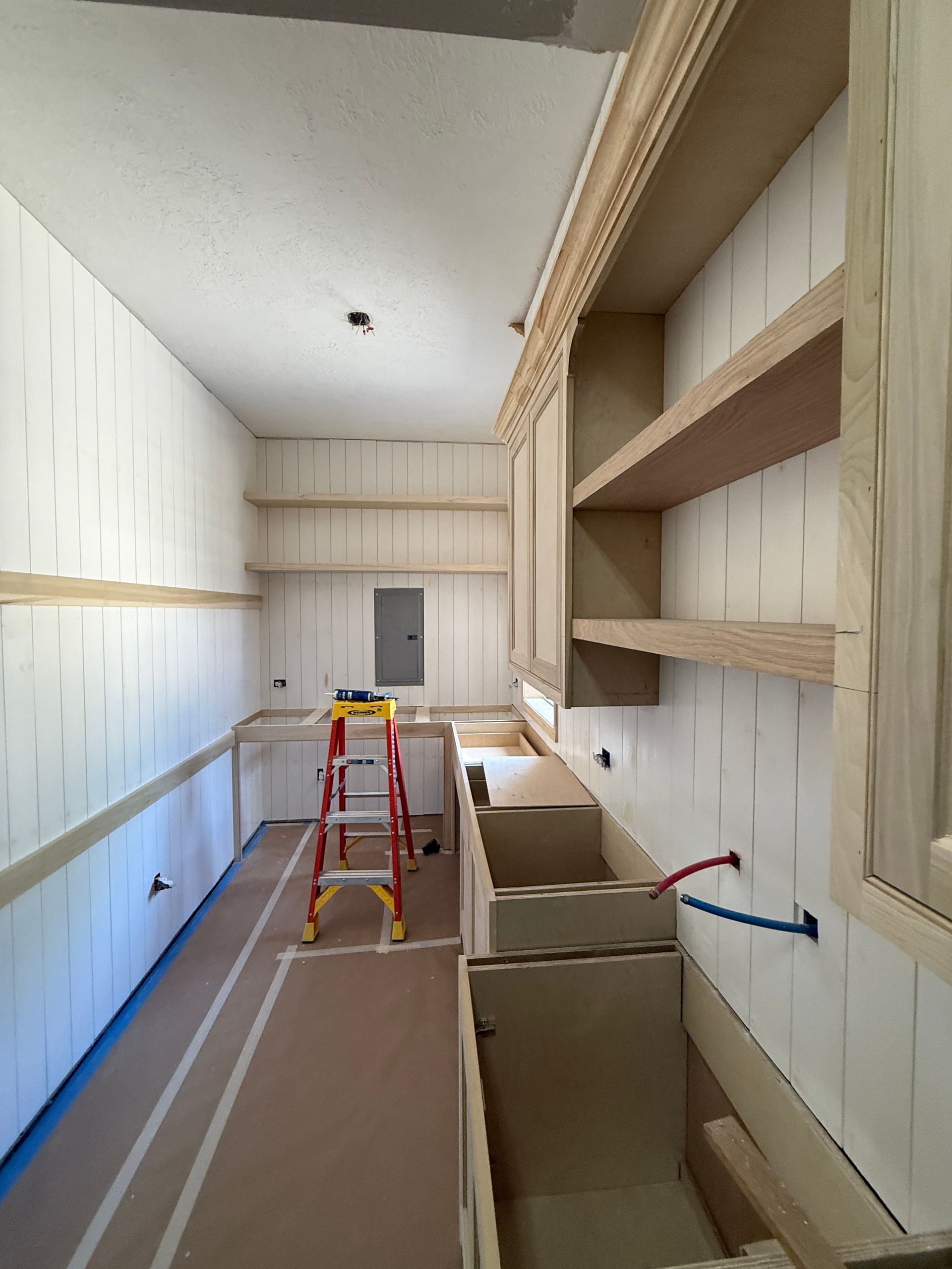 A view of a narrow, unfinished laundry room with wooden cabinetry, shelving, and an A-frame ladder on a protective floor.