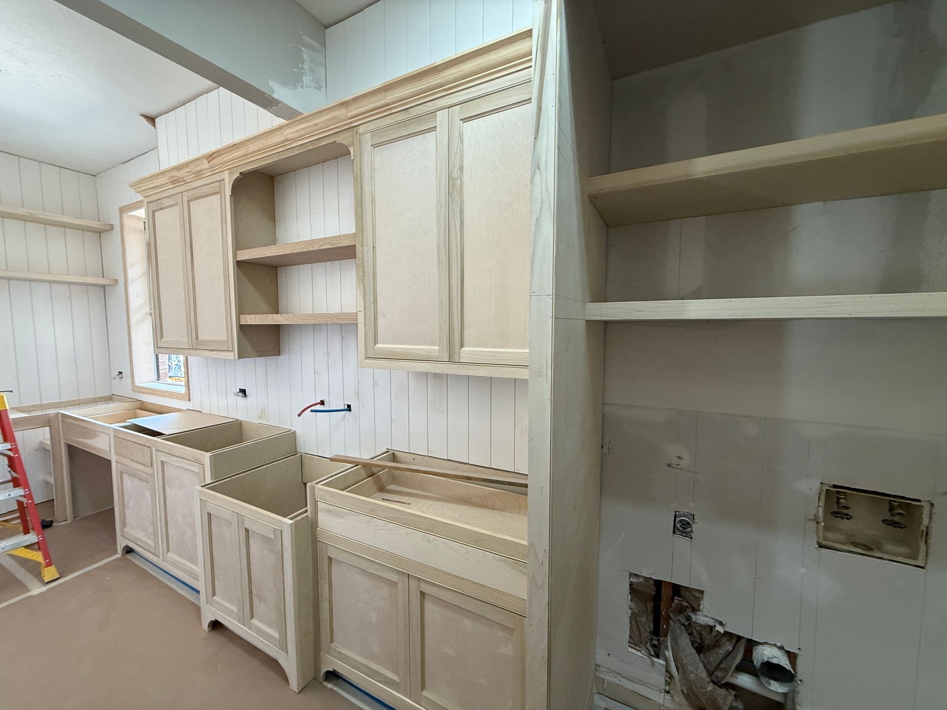 Unfinished kitchen cabinets, shelving, and a sink area under construction in a room with white paneling and exposed pipes.