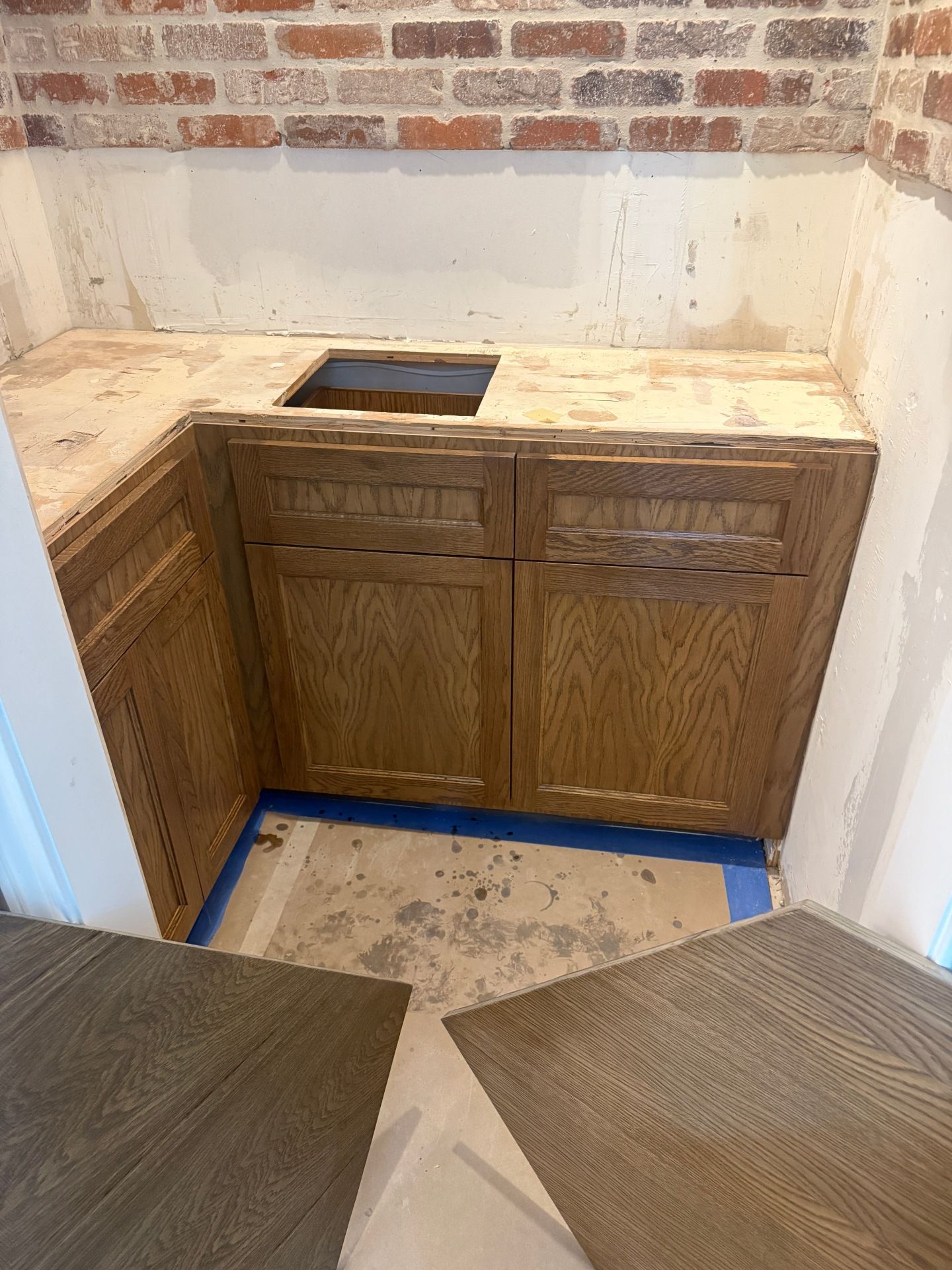L-shaped oak cabinets with a sink cutout under a brick wall during a home renovation with flooring tiles in the foreground.