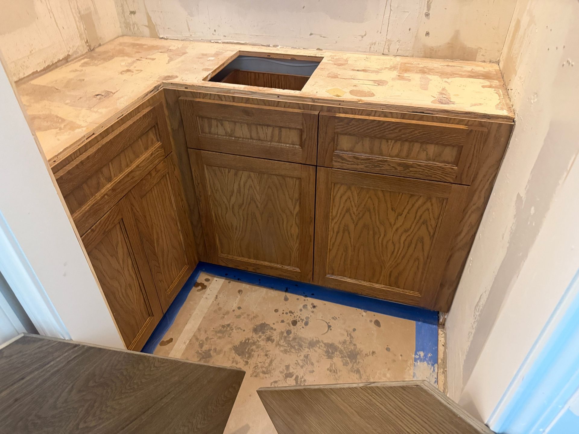 Corner oak cabinets with a countertop cutout, mid-renovation with unfinished flooring and walls in a small room.