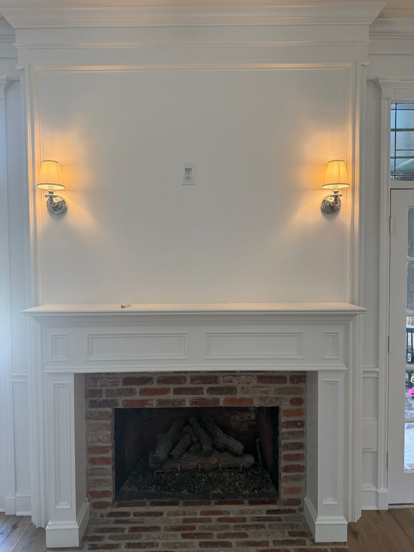 A white fireplace mantel with a brick surround, featuring two wall sconces illuminating a blank wall space above.