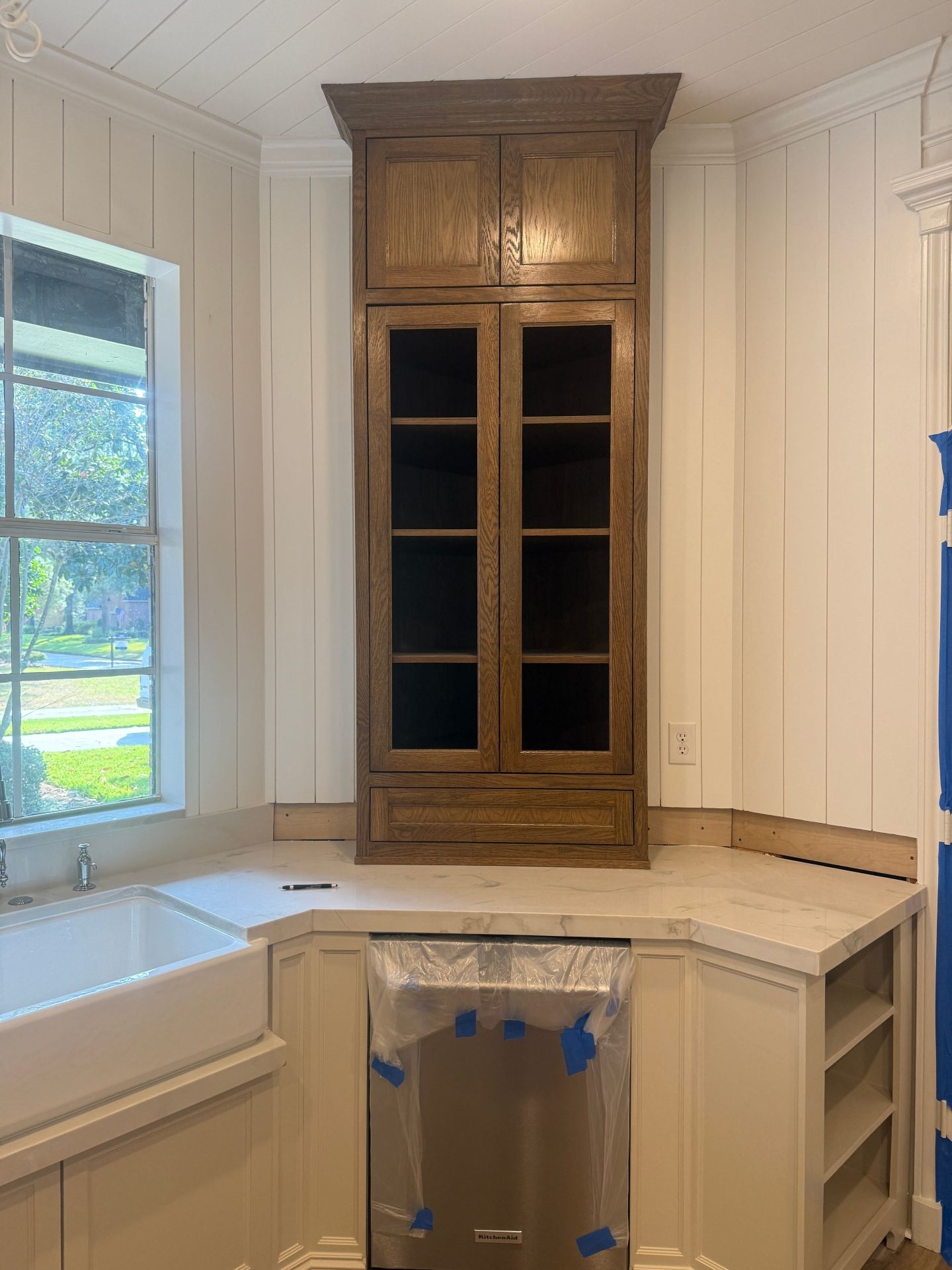 A kitchen corner with a dark wood cabinet above a white counter, a farmhouse sink, and an unfinished dishwasher.