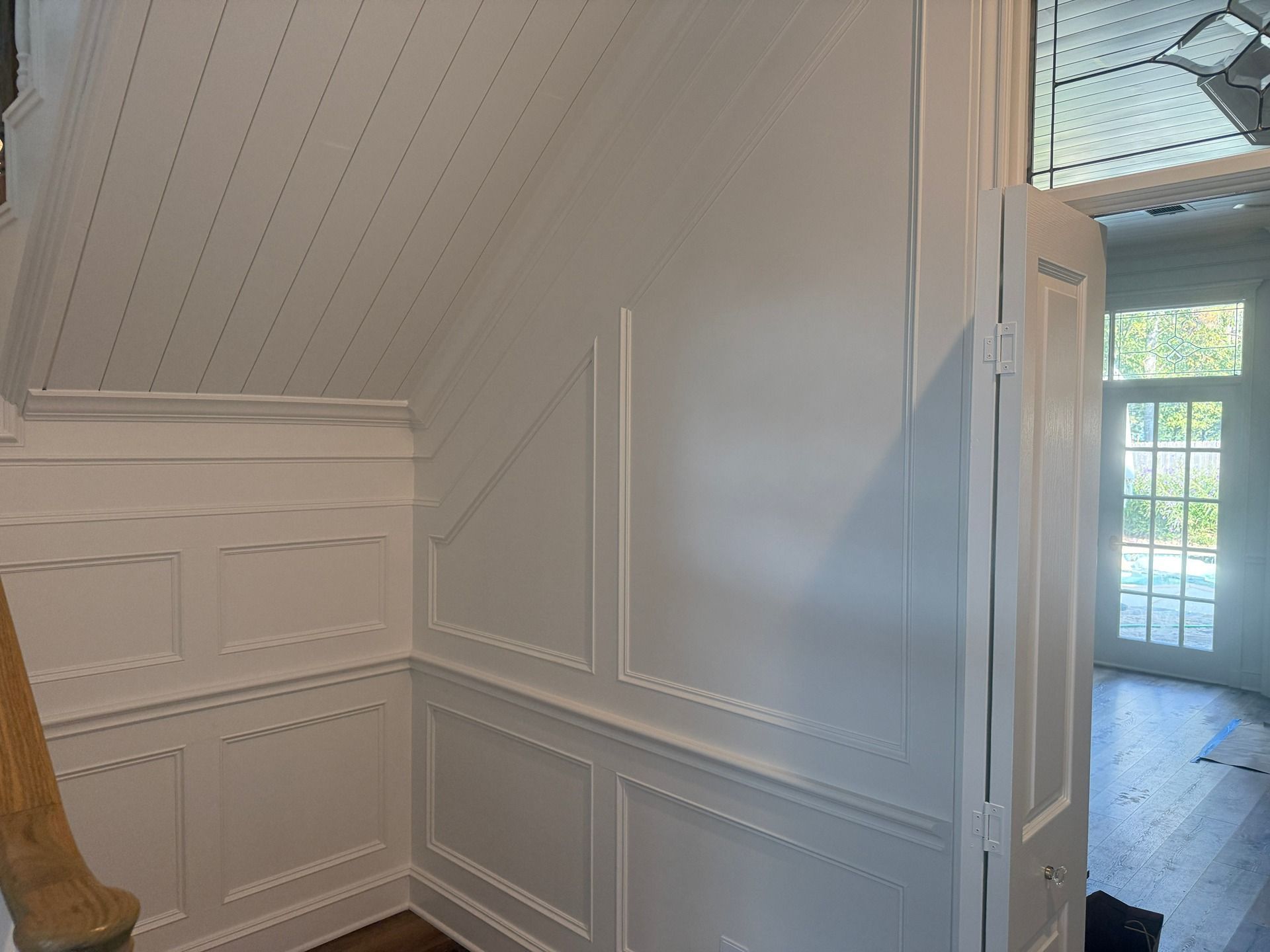 A white staircase wall featuring decorative wainscoting and paneled molding, leading toward a brightly lit glass doorway.