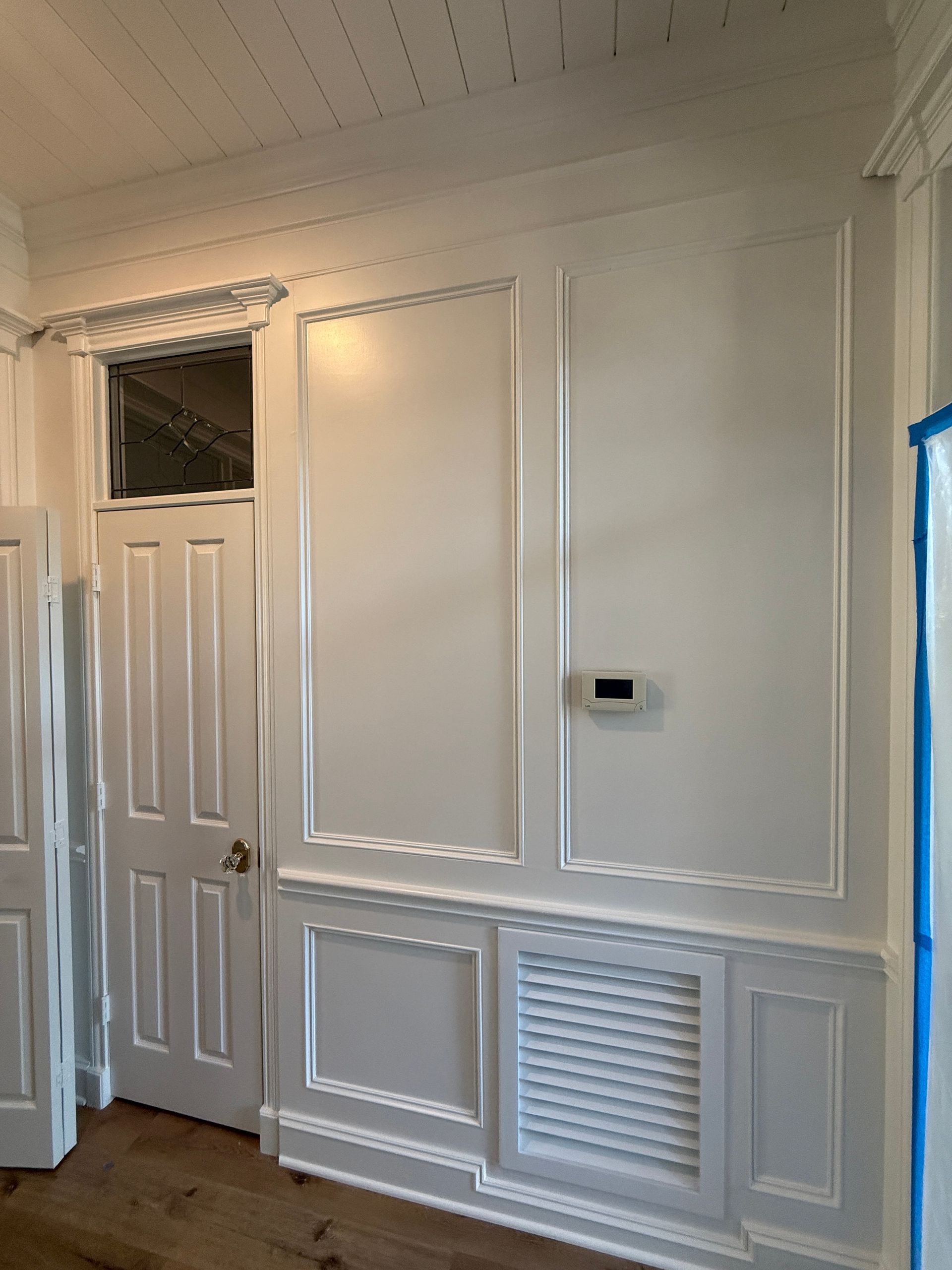 A white wall featuring decorative wainscoting, molding panels, a white wooden door, and a thermostat.