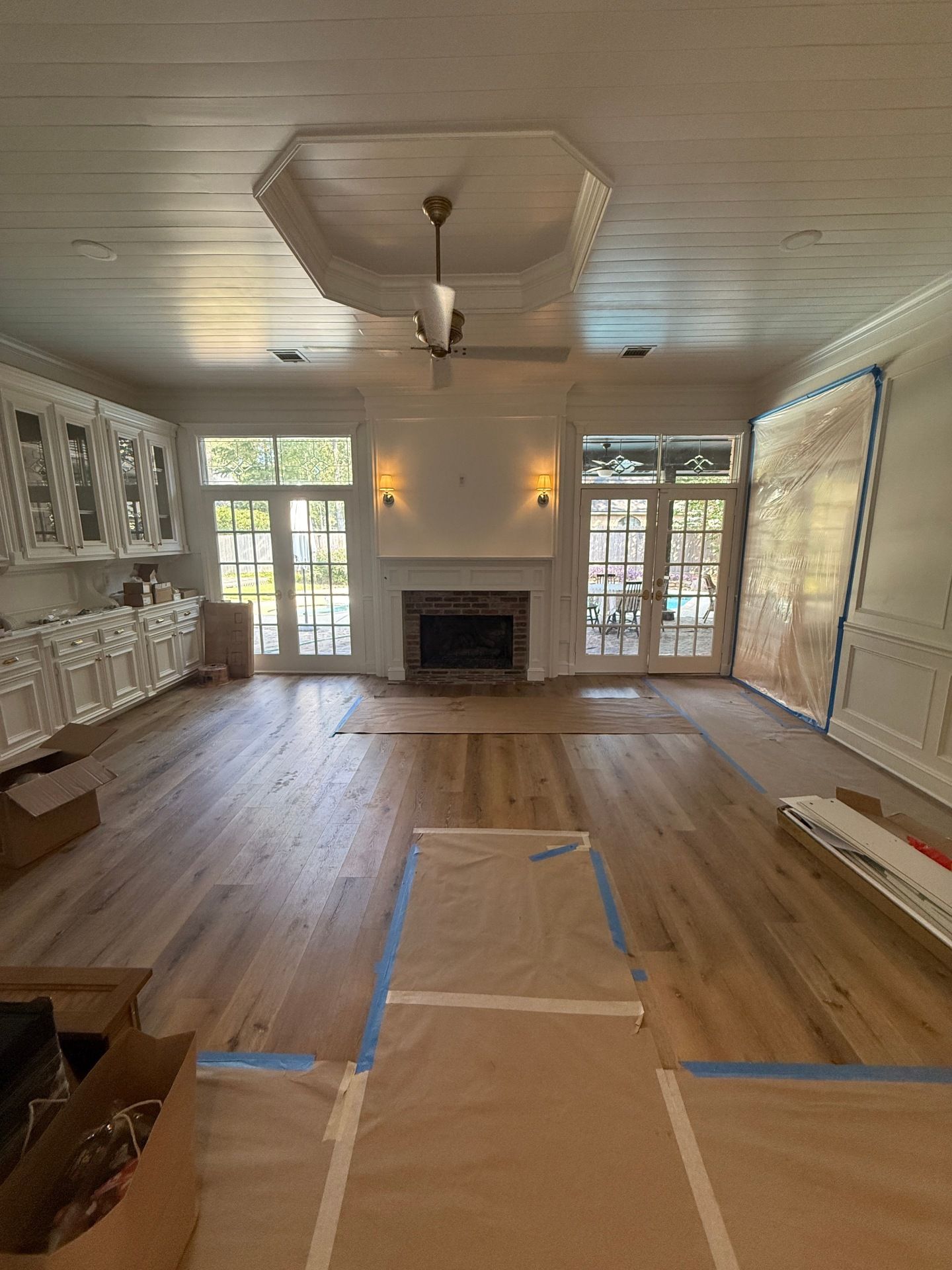A sunlit living room under renovation with new light wood flooring, a fireplace, white cabinetry, and a tray ceiling.