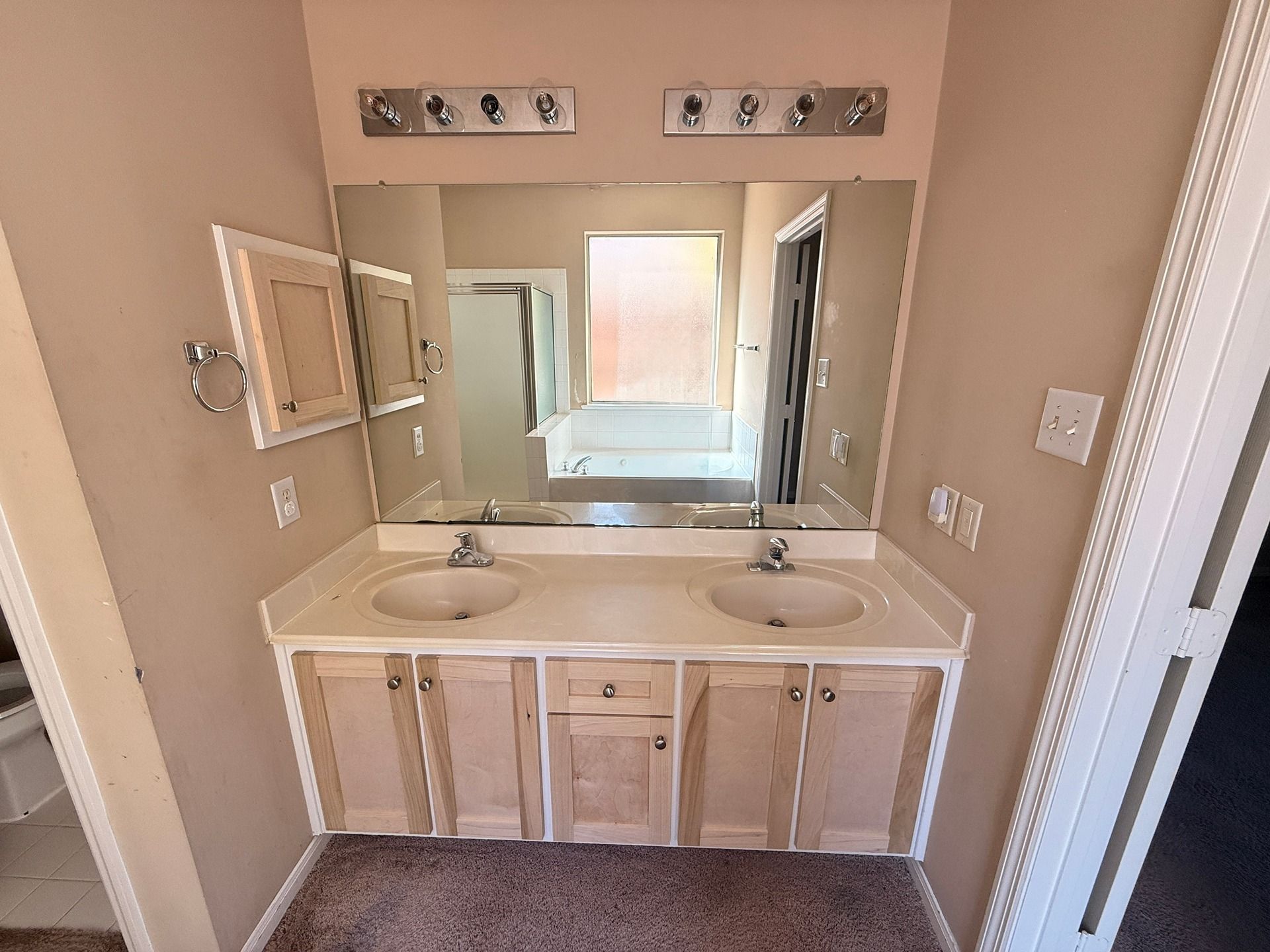 A bathroom vanity with a double sink, white countertop, light wood cabinets, two large mirrors, and two light fixtures.