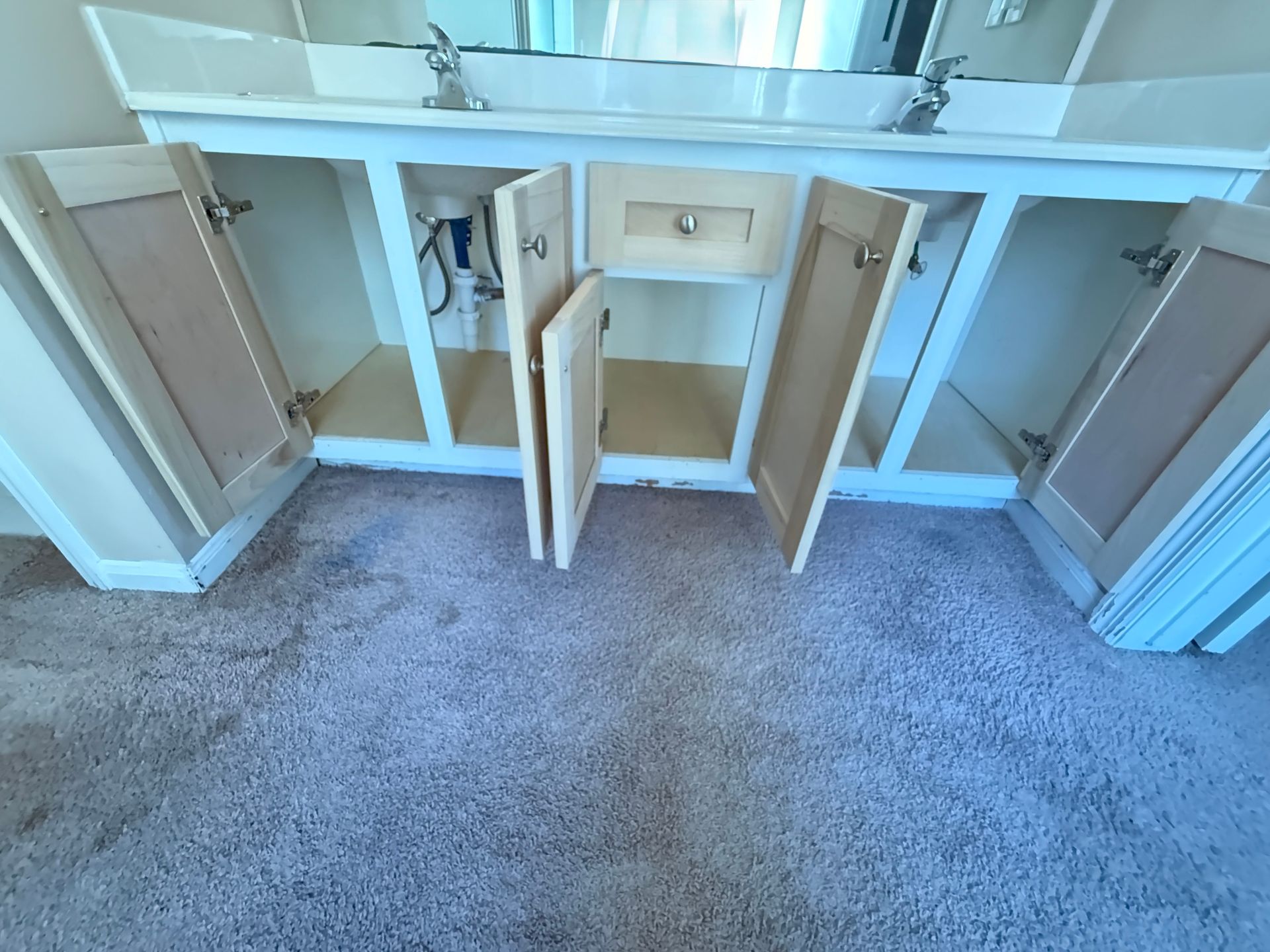 Light-colored double vanity with open cabinet doors and exposed plumbing, set on gray carpet.