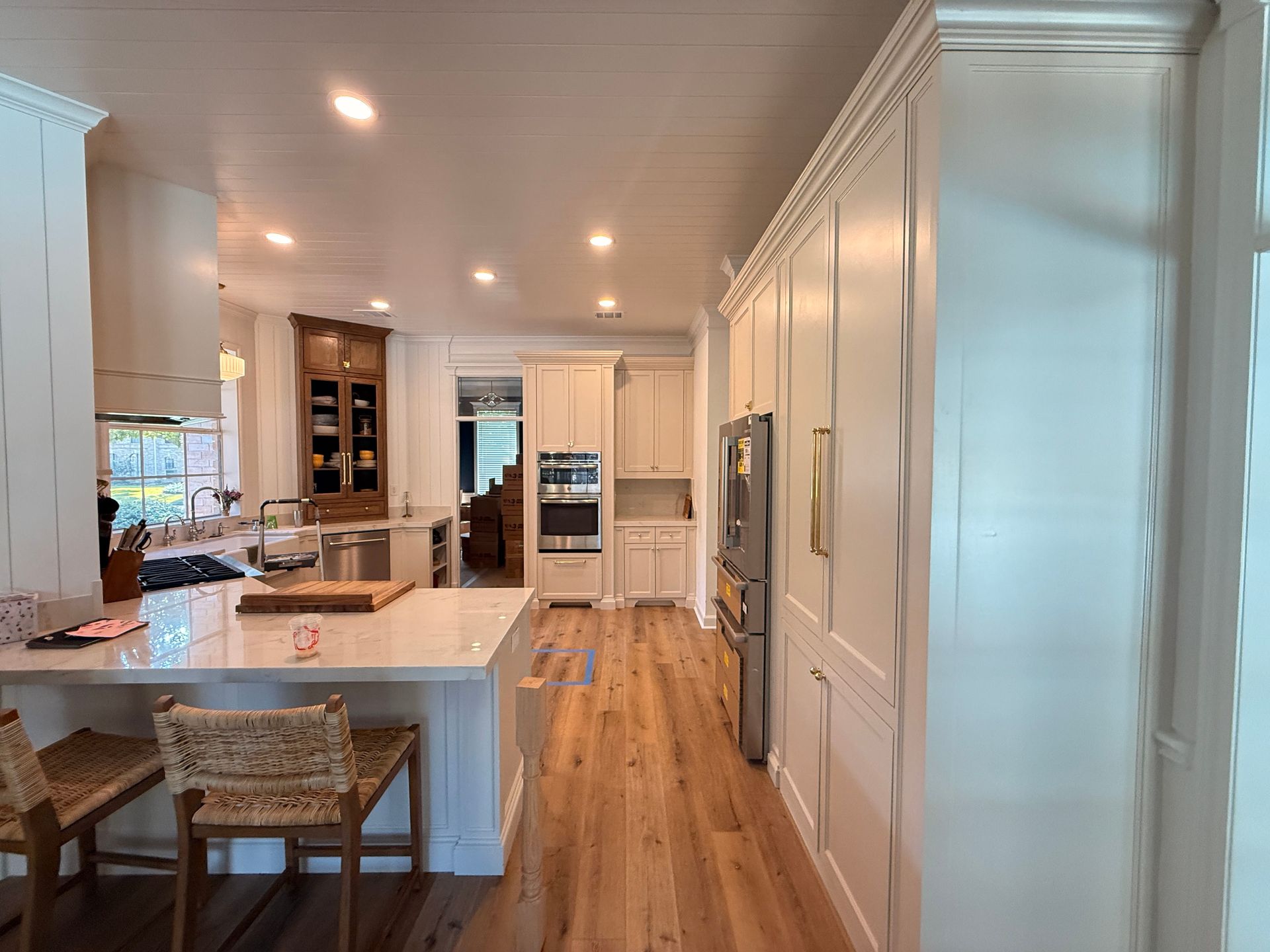 A white kitchen with a large island, wood flooring, stainless steel appliances, and a wooden china cabinet in the distance.