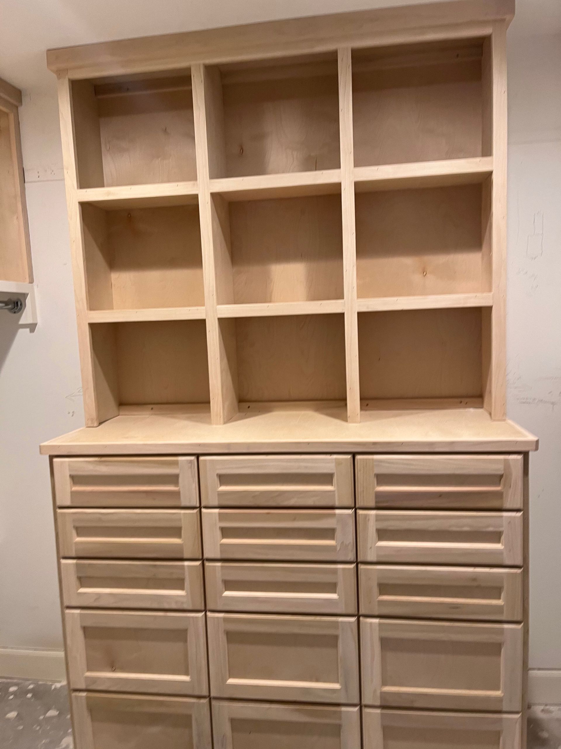 A tall, light-wood hutch with nine open storage cubbies on top and twelve matching drawers below against a white wall.