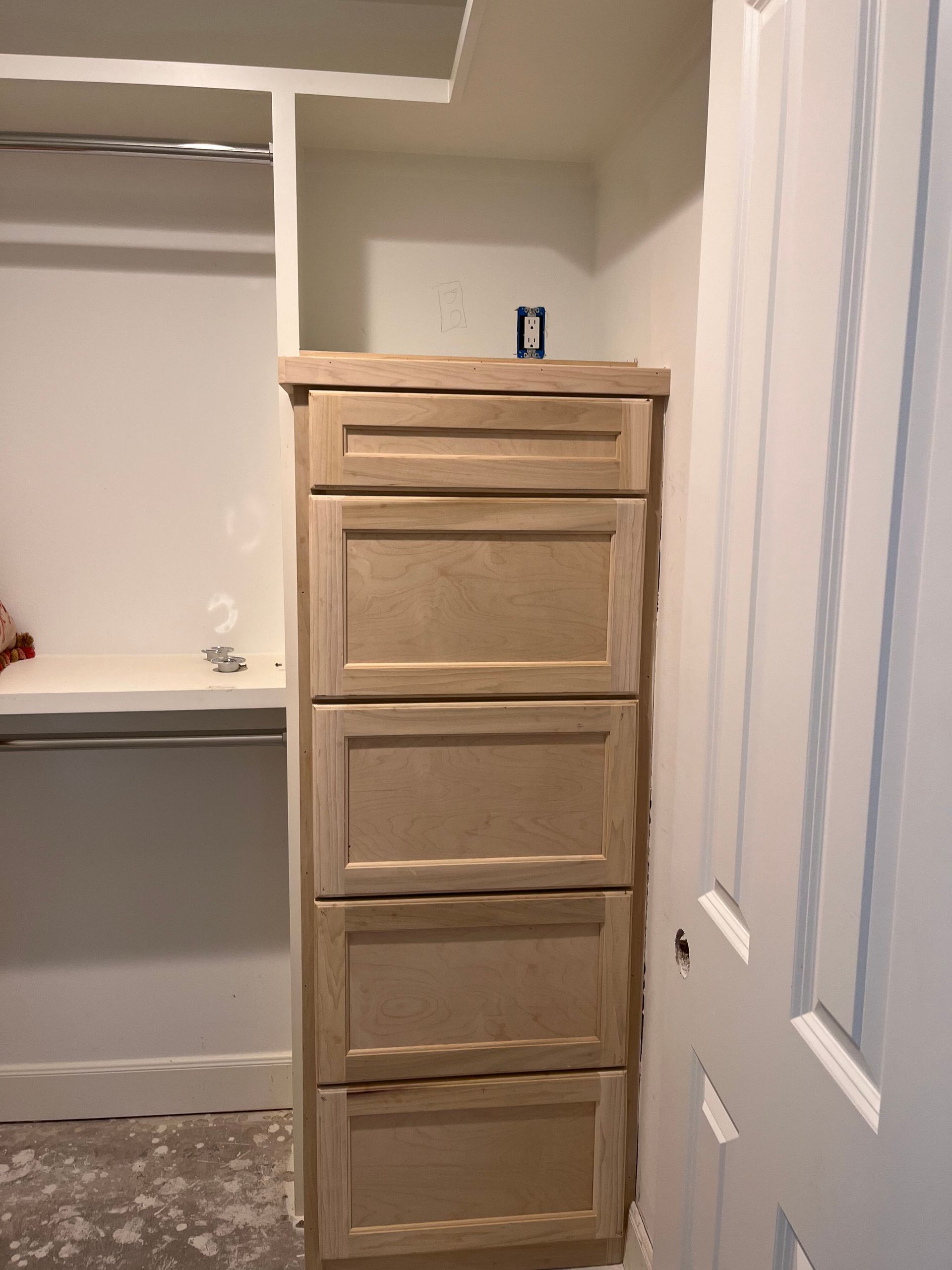 A light wood drawer unit built into a closet, positioned between a wall and a white door, with an open shelf above.